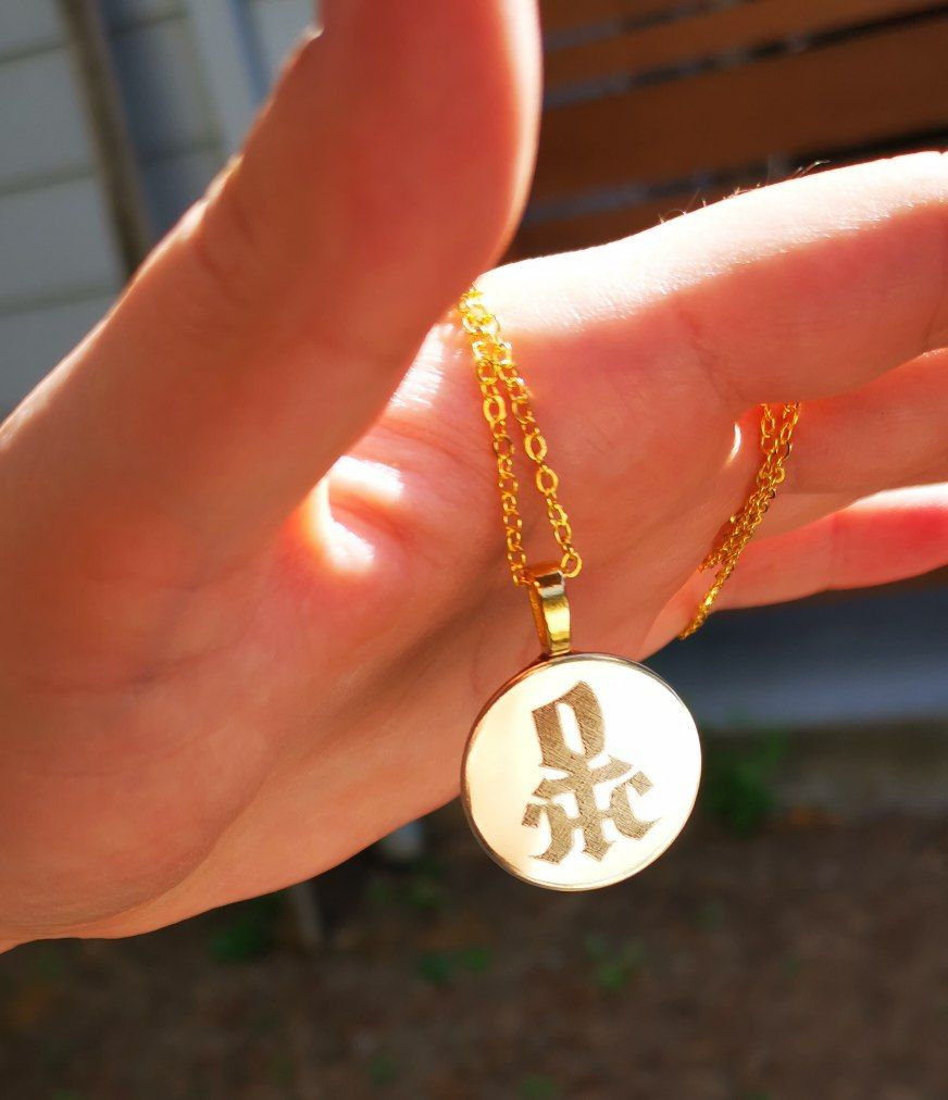 A photograph of a gold colored necklace pendant that I'm holding. It's engraved with a calligraphic astrological symbol representing a comination of Venus and Uranus (72). It's machine engraved with a tabletop Cricut machine onto a 1 inch copper disc.