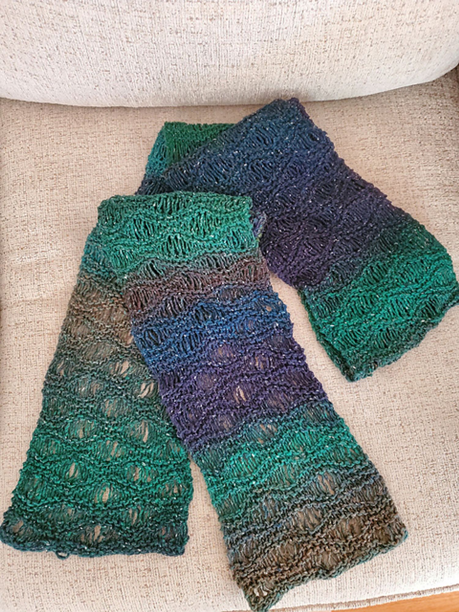 A scarf knitted in in shades of blue and greens with long slow color changes. The texture is wavy across the width of the scarf with the waves formed by drop stitches.