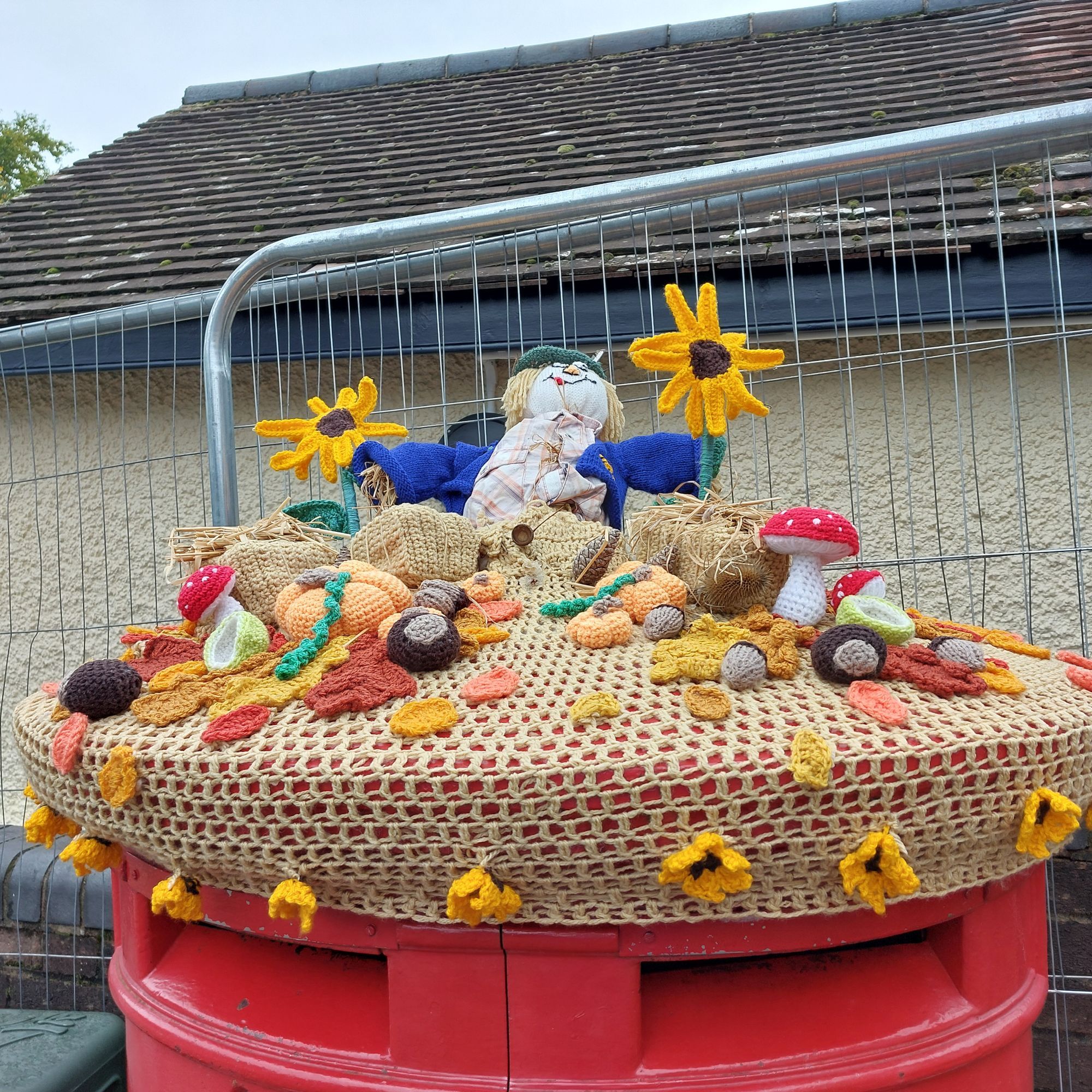 A harvest festival yarn creation with a scarecrow, sunflowers, pumpkins, autumn leaves and toadstools, on top of a postbox.