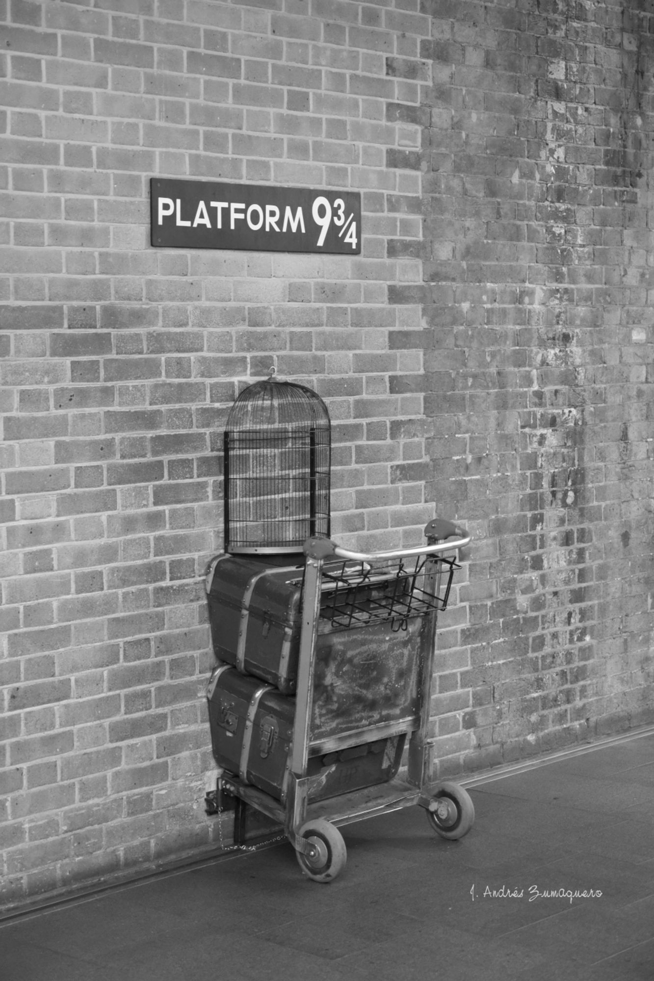 Fotografía en blanco y negro en la que hay un muro de ladrillo y un carrito, para transportar equipajes, con dos maletas grandes y una jaula. Carrito, maletas y jaula parecen estar atravesando el muro para pasar al otro lado como ocurre en la saga de películas de Harry Potter. Está en un centro comercial cerca de una estación de ferrocarril en Londres.