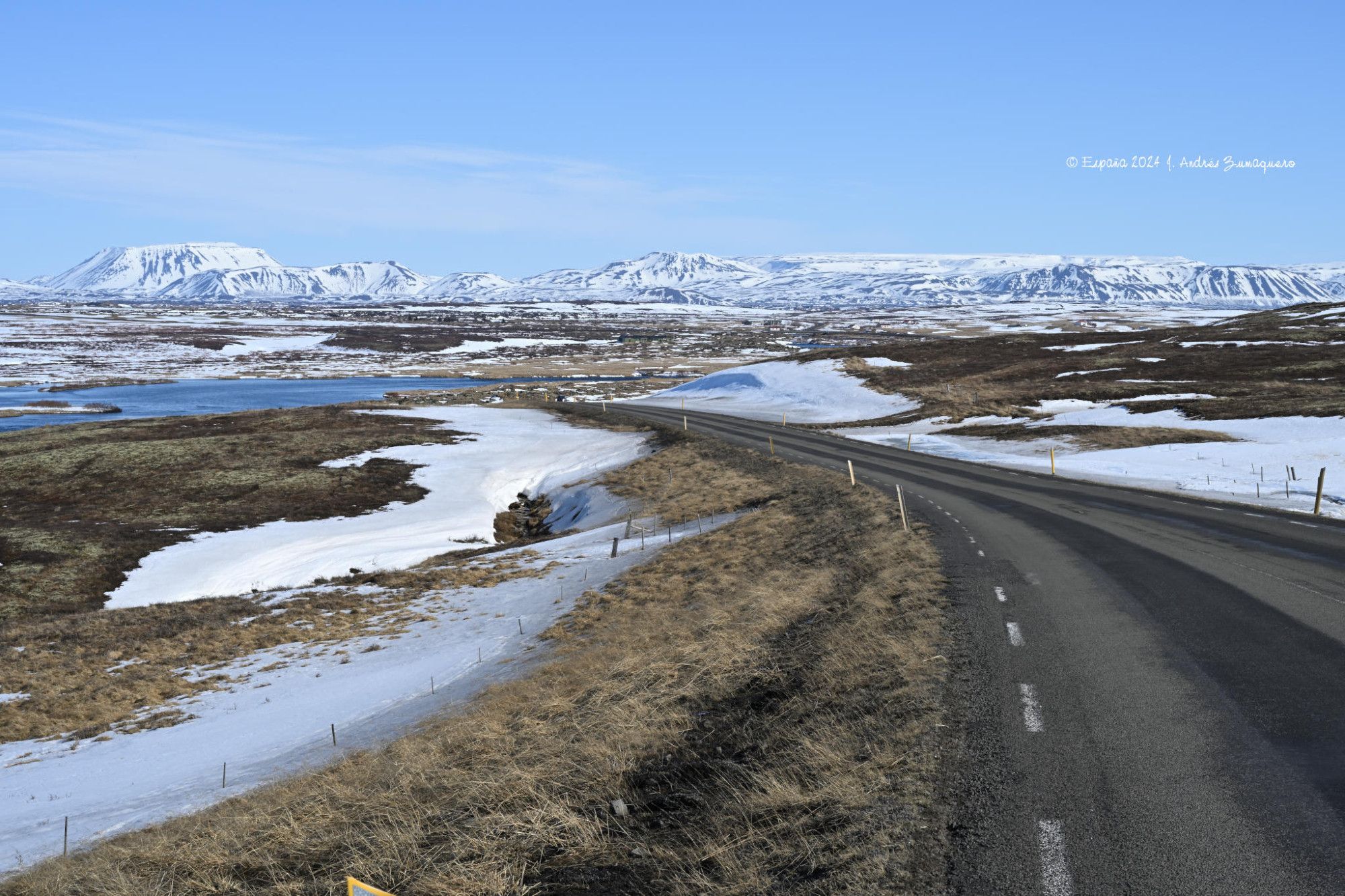 La imagen muestra unas montañas cubiertas de nieve y un cielo luminoso y celeste, en el tercio superior. Los dos tercios inferiores están formados por un paisaje con alguna nieve y también hierba seca por las heladas. La carretera R1 se ve el margen inferior derecho hasta perderse por el centro de la fotografía.