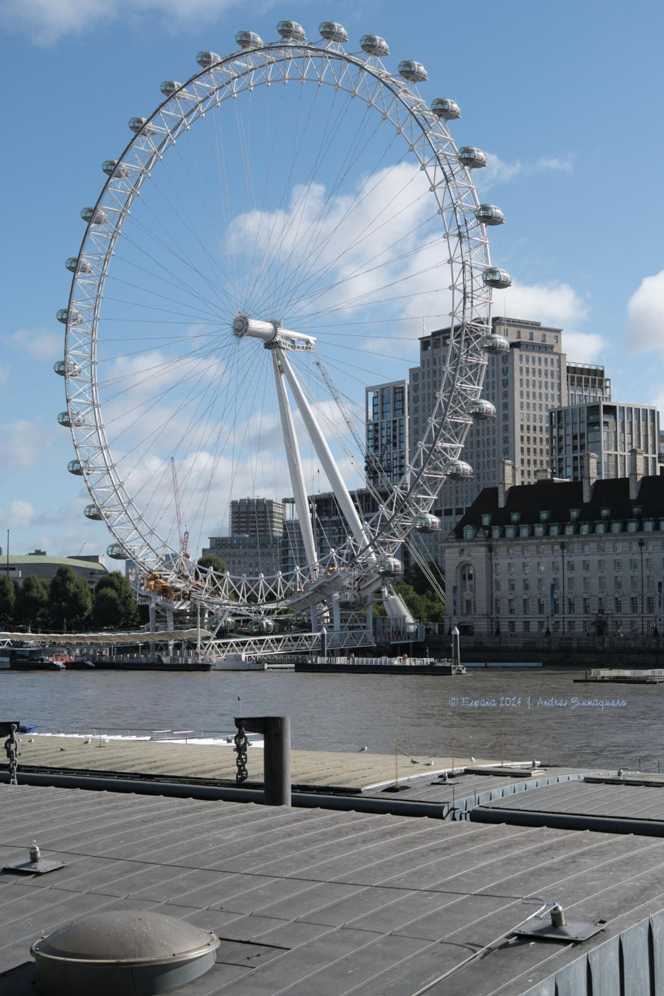 Imagen de «London Eye», la noria que se encuentra a uno de los lados del Támesis fotografiada desde el lado opuesto del río. En la foto aparece también un tramo del río y algunos edificios altos tras la noria. El cielo es de color celeste con algunas nubes.