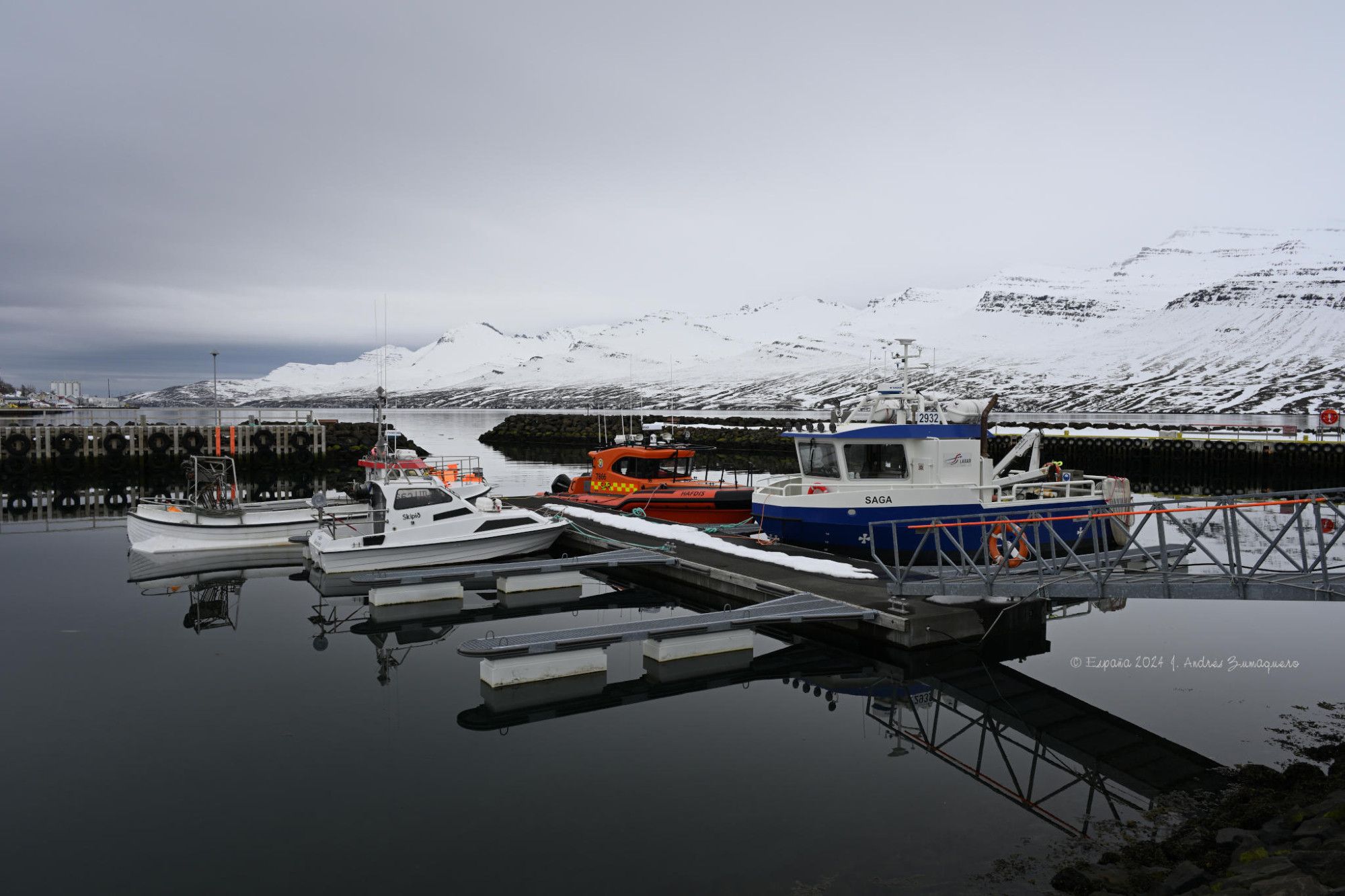 La mitad inferior de la fotografía muestra unos embarcaderos y varios barcos atracados en ellos. El agua está completamente cristalina, de un  color gris oscuro casi negro. La mitad superior de la imagen es de un gris claro casi blanco, donde aparecen cielo y una pequeña montaña cubierta por nieve.