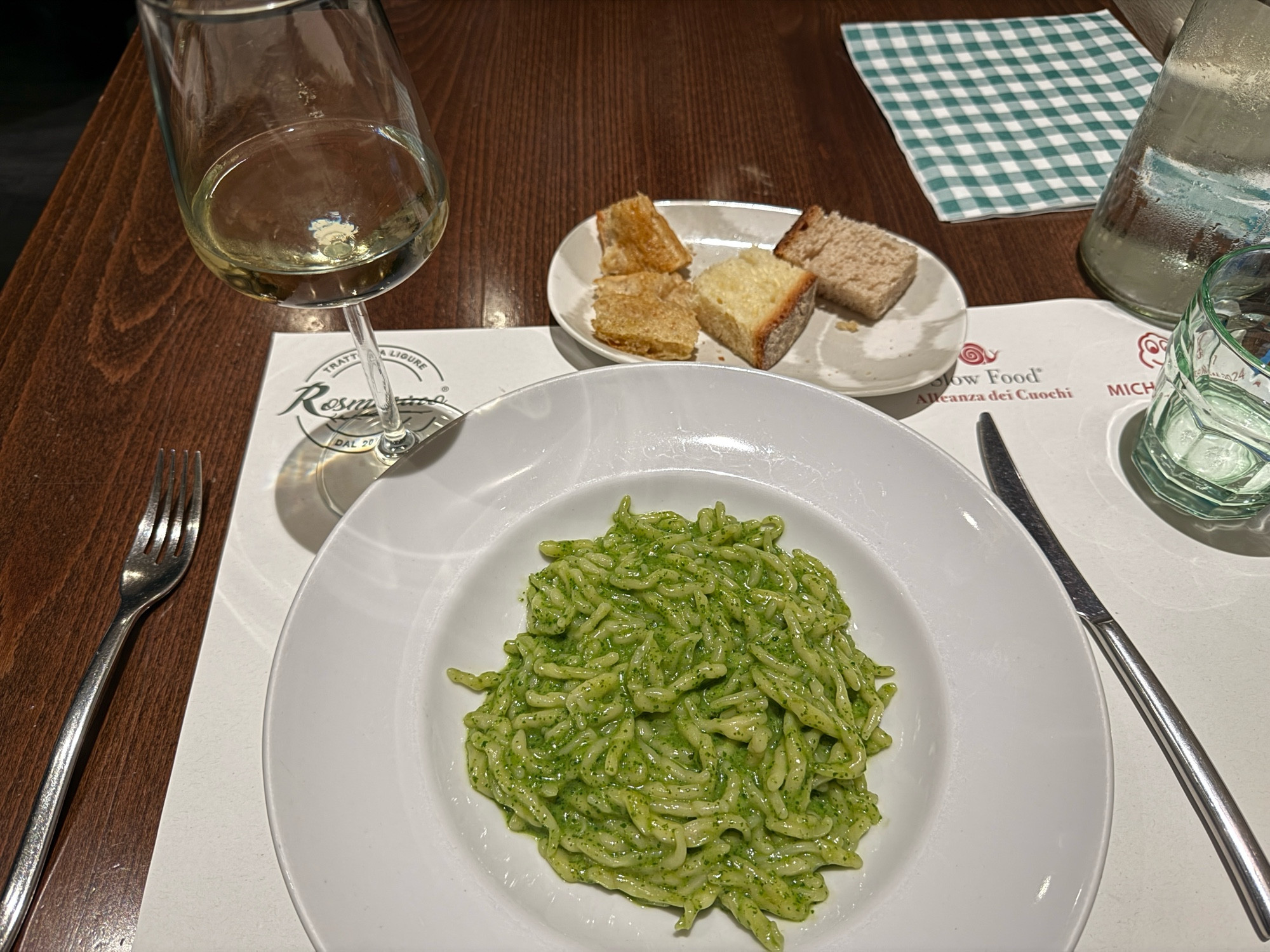 A bowl of pesto pasta with a glass of white wine and some bread and focaccia nearby