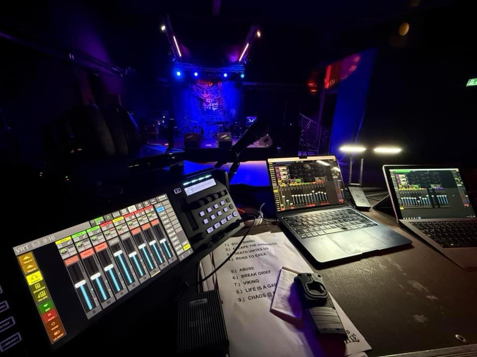 Behringer Wing Rack Mischpult links am Bildrand. Zwei Laptops zum steuern auf dem Tisch. Im Hintergrund eine dunkelblau beleuchtete Bühne kurz vor der Show. 