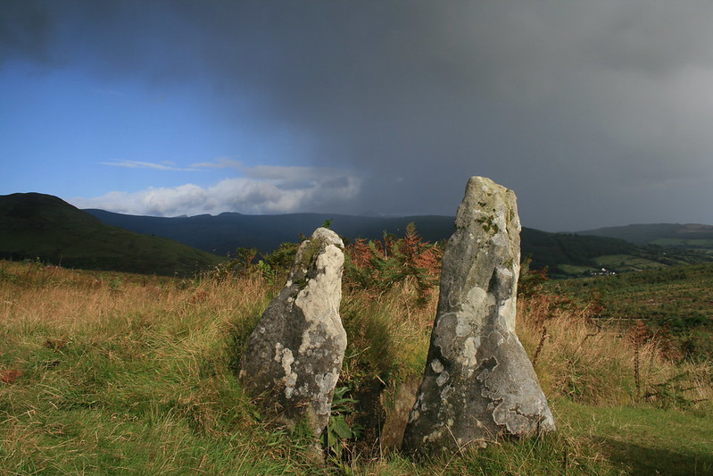 Very dark rain clouds rolling in from the righthand side of the picture, but the sky on the left is still blue and the foreground is in sunshine. Two upright stones mark the entrance to the chamber.