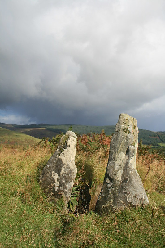 The two upright entrance stones of a very badly damaged chambered tomb. The stones are in sunlight but far beyond them the hills are dark with cloud