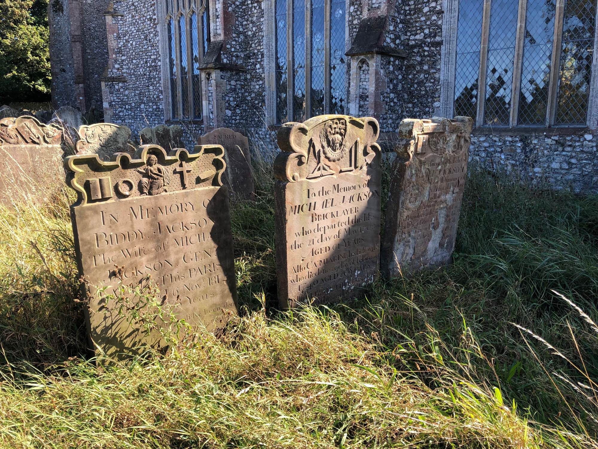 Three carved headstones in a graveyard. The middle one is for a bricklayer and has his tools carved on it