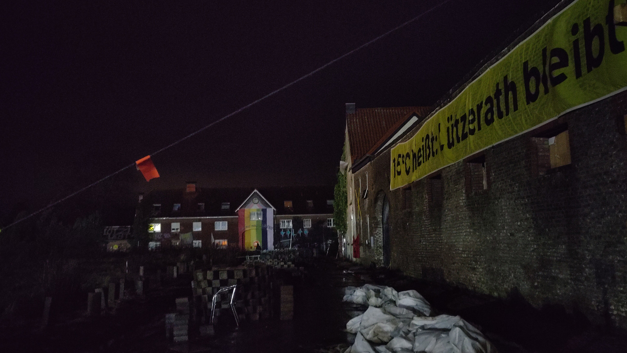 Dunkle Nacht. Rechts die Mauer des ehemaligen Wachtmeisterhofs mit Banner "1,5°C heißt Lützerath bleibt". Auf der Frontseite ein weiterer Hof, angestrahlt mit vertikalen Streifen in Regenbogenfarben.