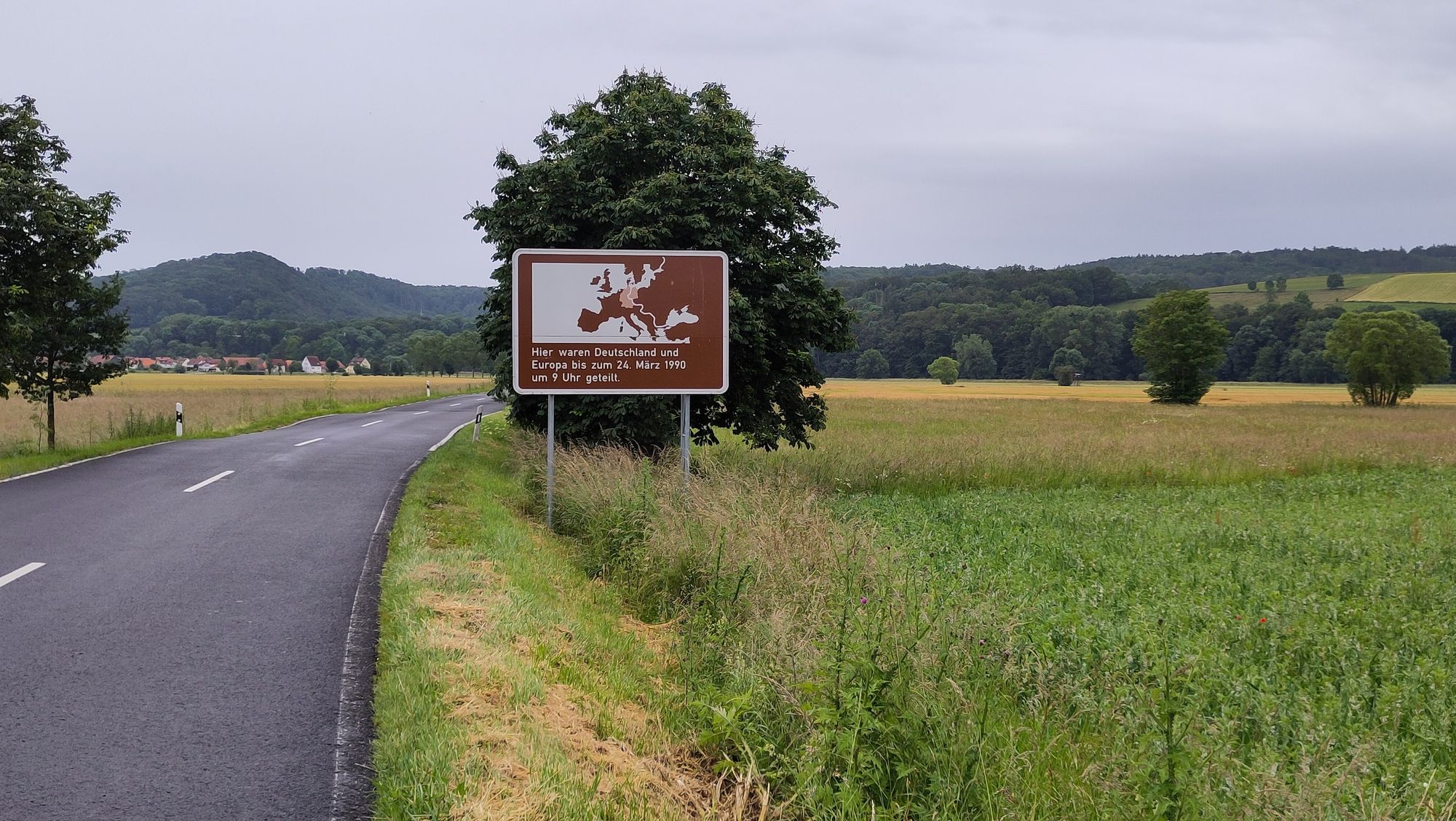 Landschaft mit Straße, Landwirtschaft, Bäumen und einem großen Schild mit Europakarte und der Aufschrift "Hier waren Deutschland und Europa bis zum 24. März 1990 um 9 Uhr geteilt". Im Hintergrund Berge und ein Dorf.