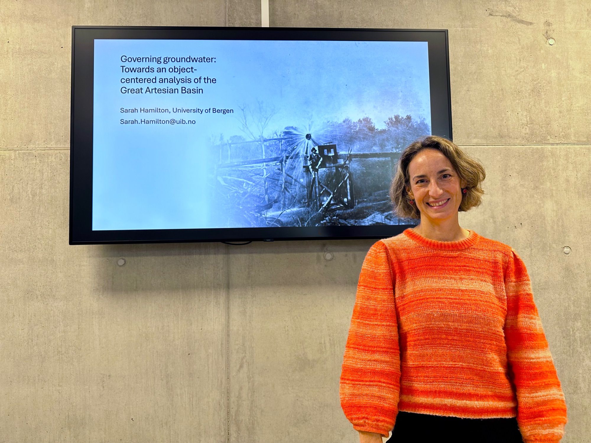 Sarah Hamilton in a brightly colored sweater in front of a monitor with a picture of an old irrigation scene with the text "Governing Groundwater: Towards an object-centered analysis of the Great Artesian Basin"