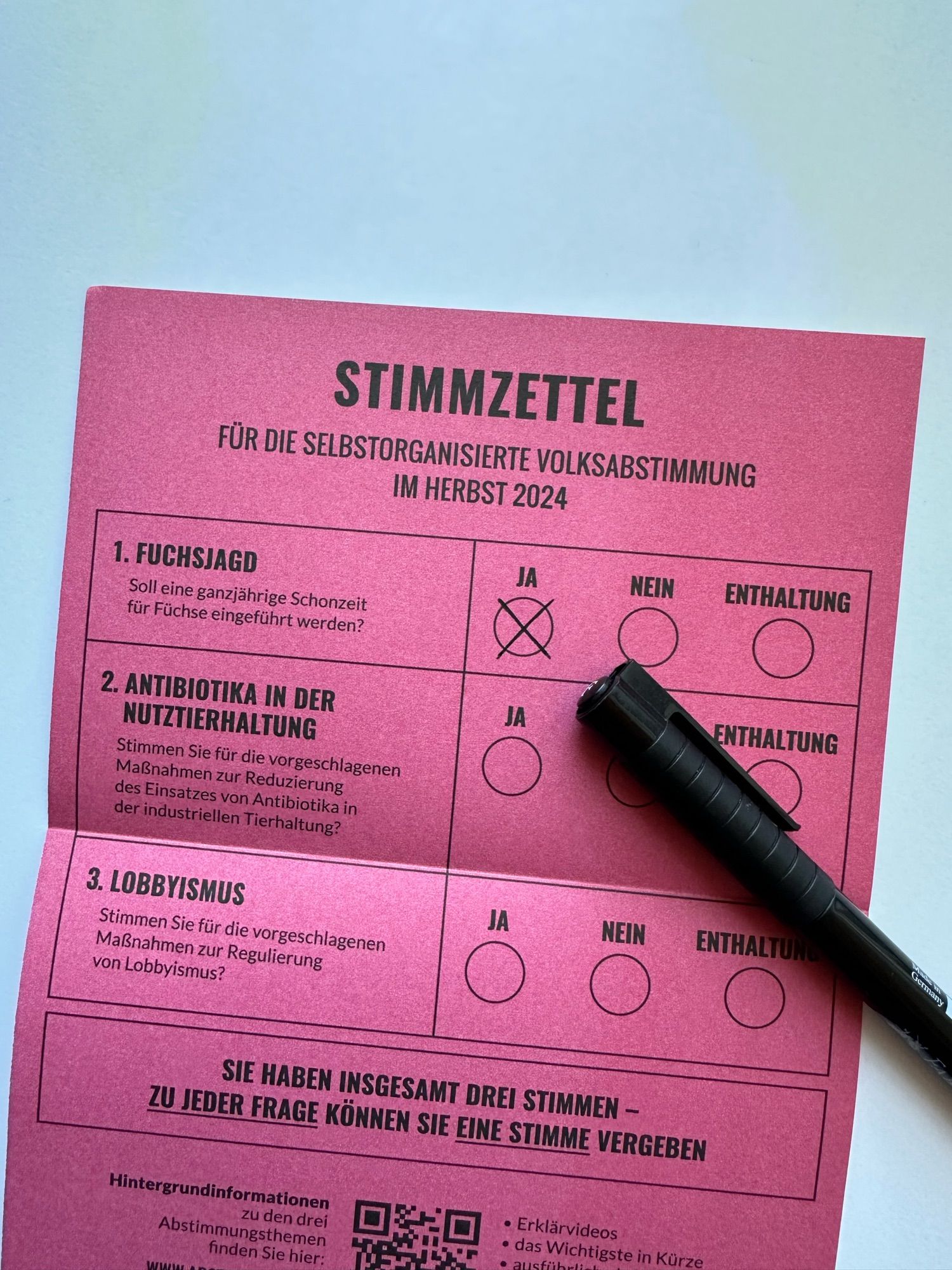 Das Bild zeigt einen rosa Stimmzettel für eine selbstorganisierte Volksabstimmung im Herbst 2024. Es gibt drei Themen, zu denen abgestimmt werden soll:

1. **Fuchsjagd**: "Soll eine ganzjährige Schonzeit für Füchse eingeführt werden?" Hier wurde mit "Ja" gestimmt.
2. **Antibiotika in der Nutztierhaltung**: "Stimmen Sie für die vorgeschlagenen Maßnahmen zur Reduzierung des Einsatzes von Antibiotika in der industriellen Tierhaltung?" Hier wurde noch keine Stimme abgegeben.
3. **Lobbyismus**: "Stimmen Sie für die vorgeschlagenen Maßnahmen zur Regulierung von Lobbyismus?" Auch hier wurde noch keine Stimme abgegeben.

Am unteren Rand gibt es Hinweise zur Abstimmung sowie einen QR-Code, der vermutlich weiterführende Informationen bietet. Ein schwarzer Stift liegt auf dem Stimmzettel.