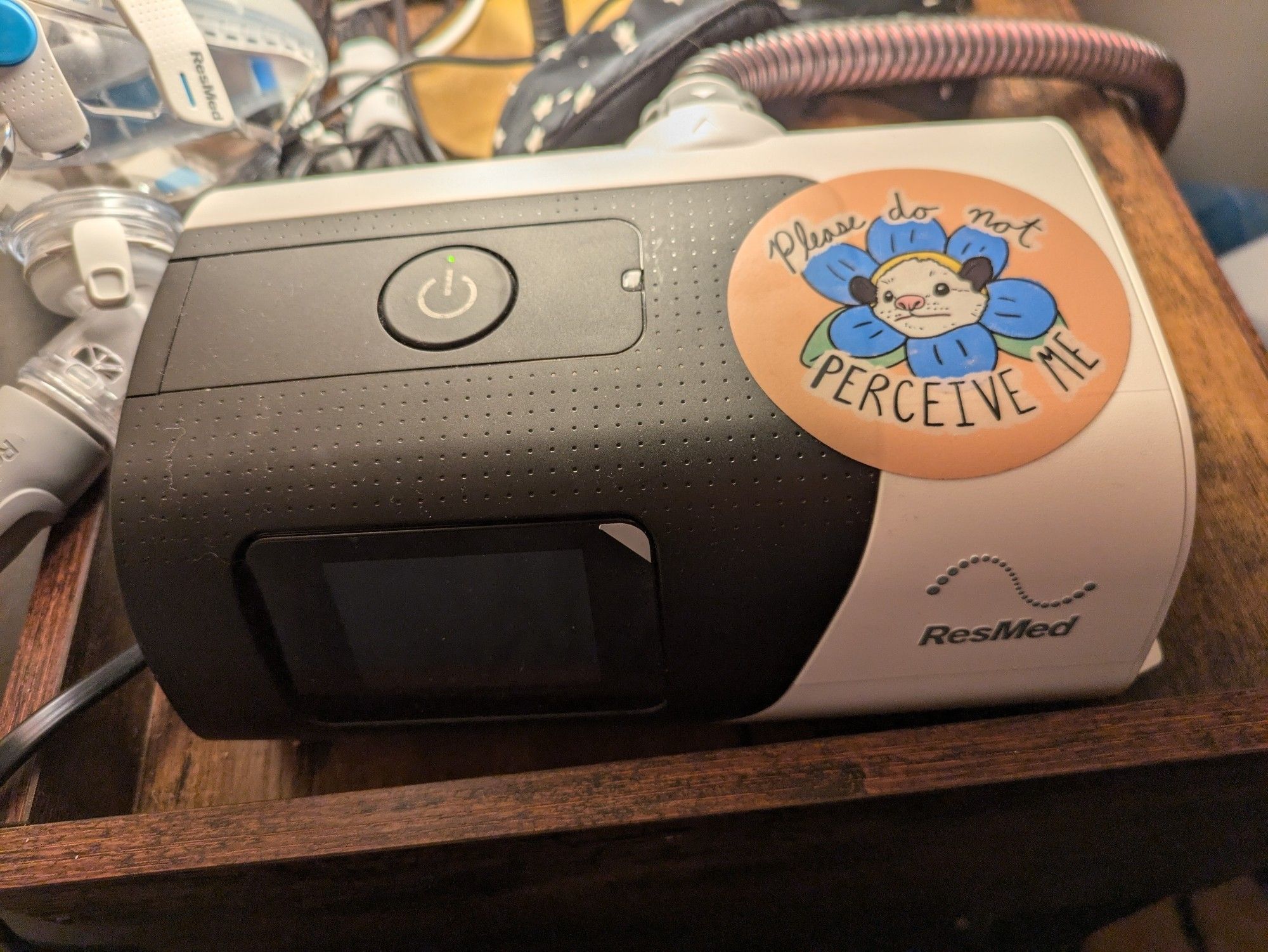 A CPAP device with a sticker on it. The sticker is an opossum's face, surrounded by flower petals, and text reading, please do not perceive me