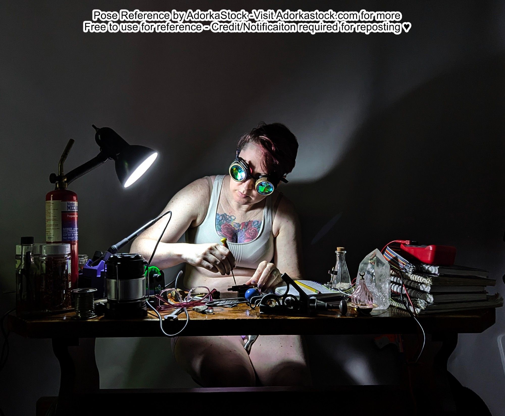 Portrait of a model sitting at a bench table that's covered in tools and supplies. She's using a screwdriver on something in the clutter and wearing silly goggles. There's a strong, bright light on the table illuminating her hands and casting harsh shadows.