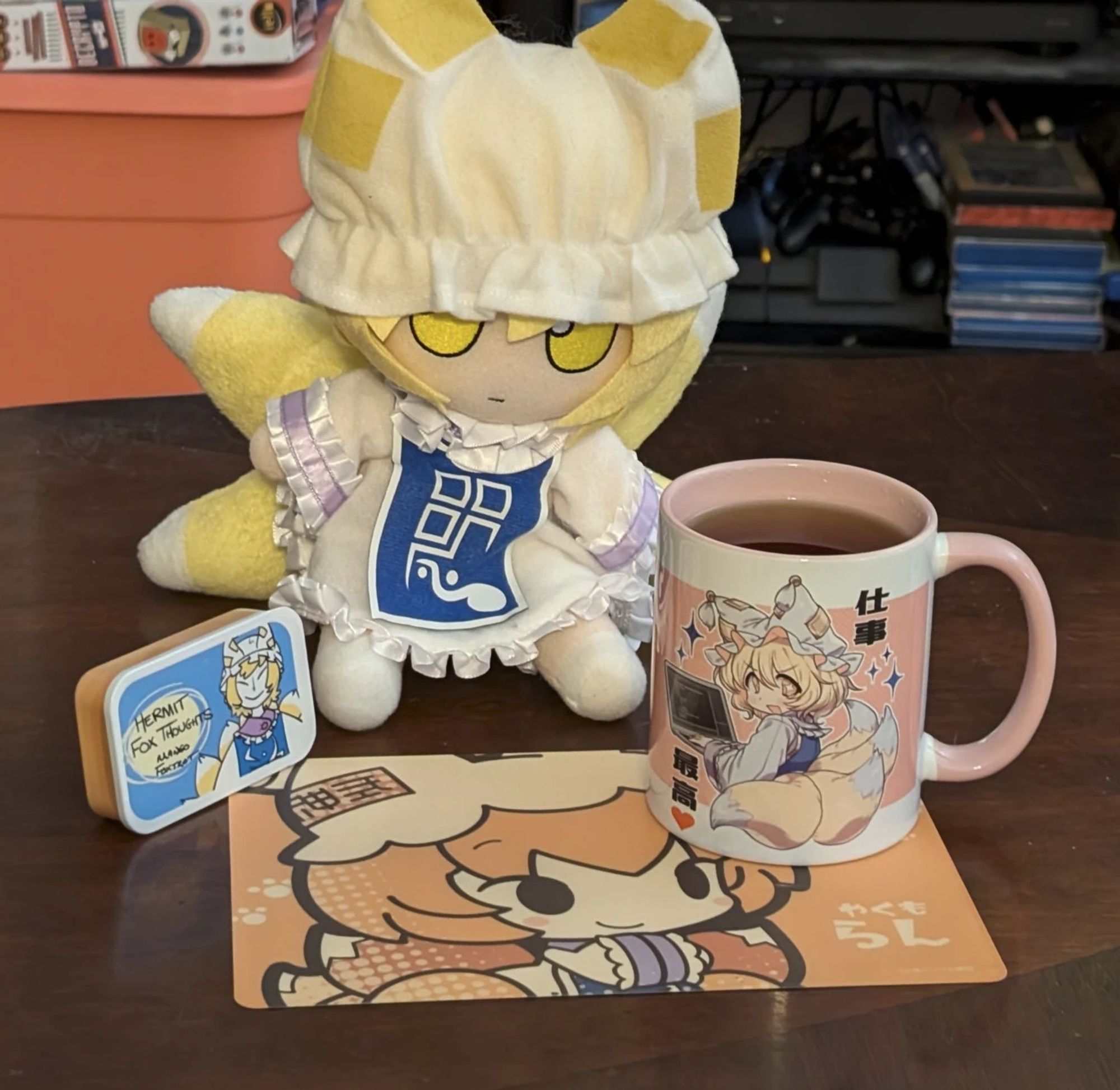 A photo of a mug of tea on a table. The mug has an illustration of Ran Yakumo, nine-tailed fox from Touhou Project, on it, and it's sitting on a mousepad with Ran, and it's the two of them are in front of a Ran fumo plushie, which is sitting next to a sample tin of tea labeled "Hermit Fox Thoughts," which, as you might have guessed, has an illustration of Ran on it.