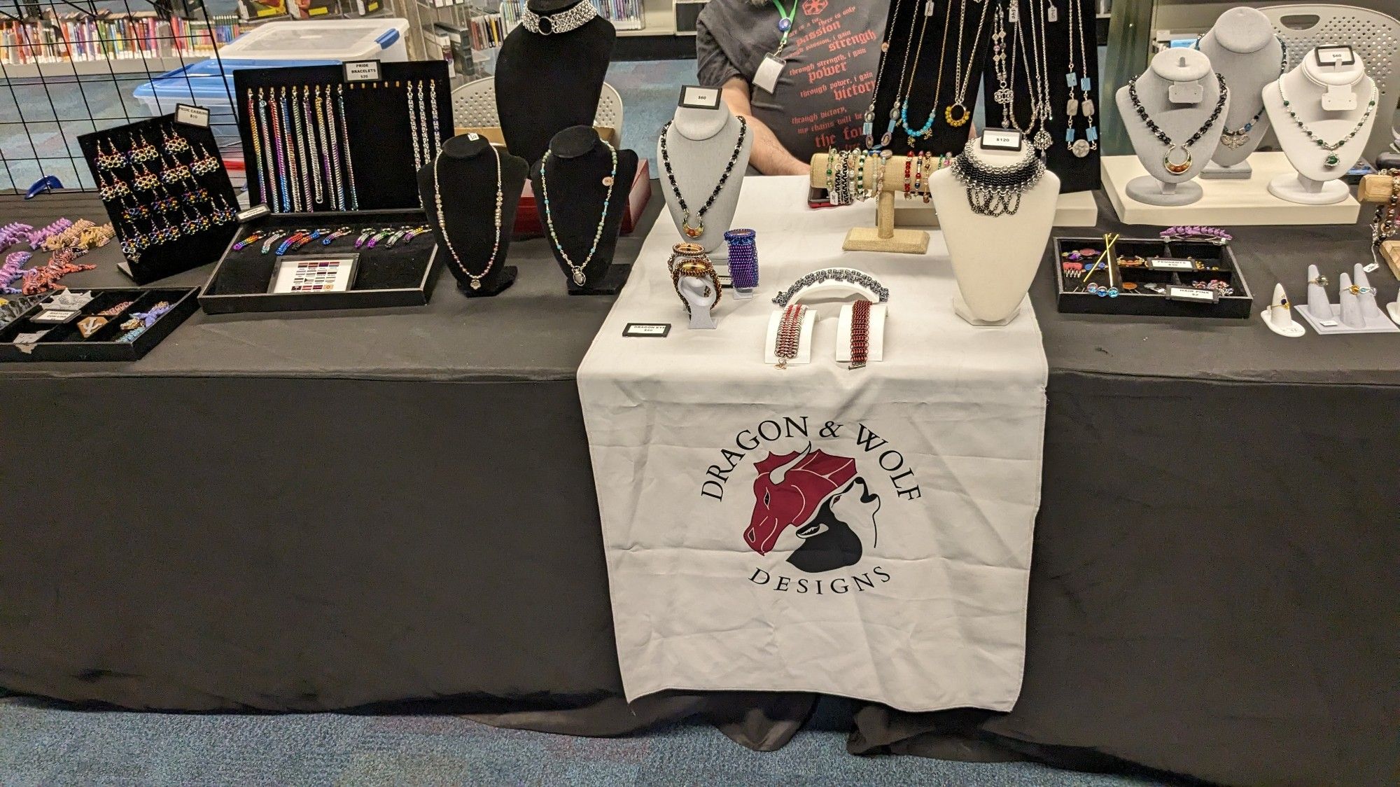 Table display with jewelry & 3D prints. A white banner hangs in front of the table with the logo for Dragon & Wolf Designs