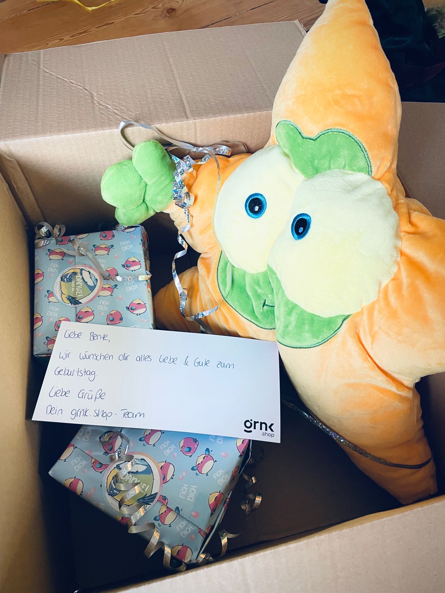 Foto in ein offenes Paket, man sieht eine Karte mit Grüßen zum Geburtstag, zwei verpackte Geschenke und ein großes Plüschtier. Nämlich das Gronkh Maskottchen den Lurch in einem Sternkostüm.