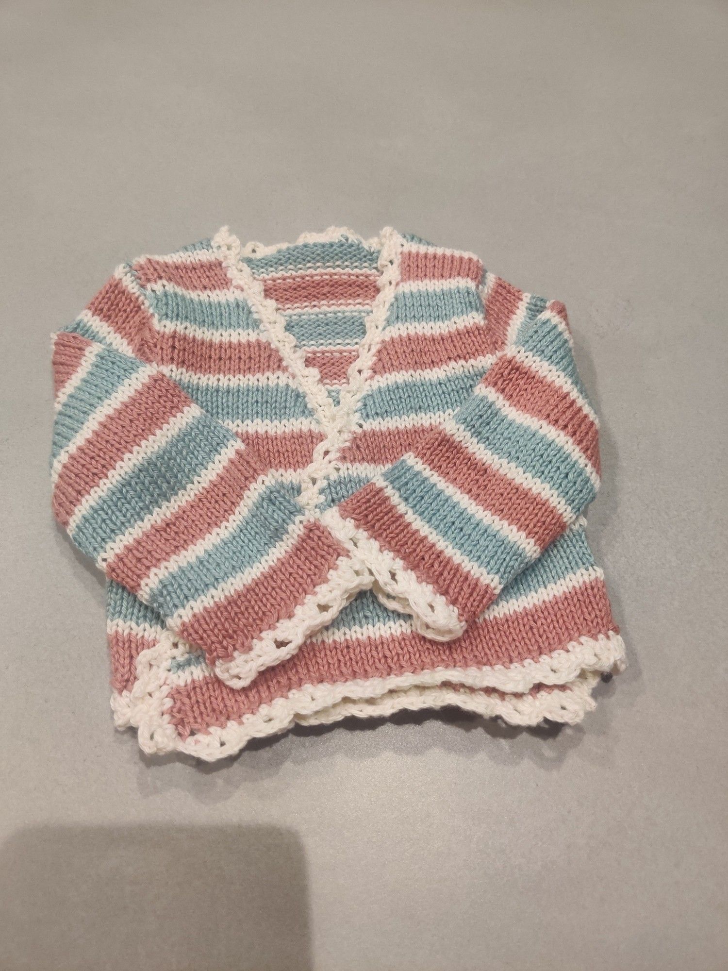 Een gebreid vestje voor een baby. Het vestje heeft mintgroene en oudroze strepen met daar tussen een rijtje wit. Om de randjes zitten gehaakte biesjes die een soort kanten randje maken.