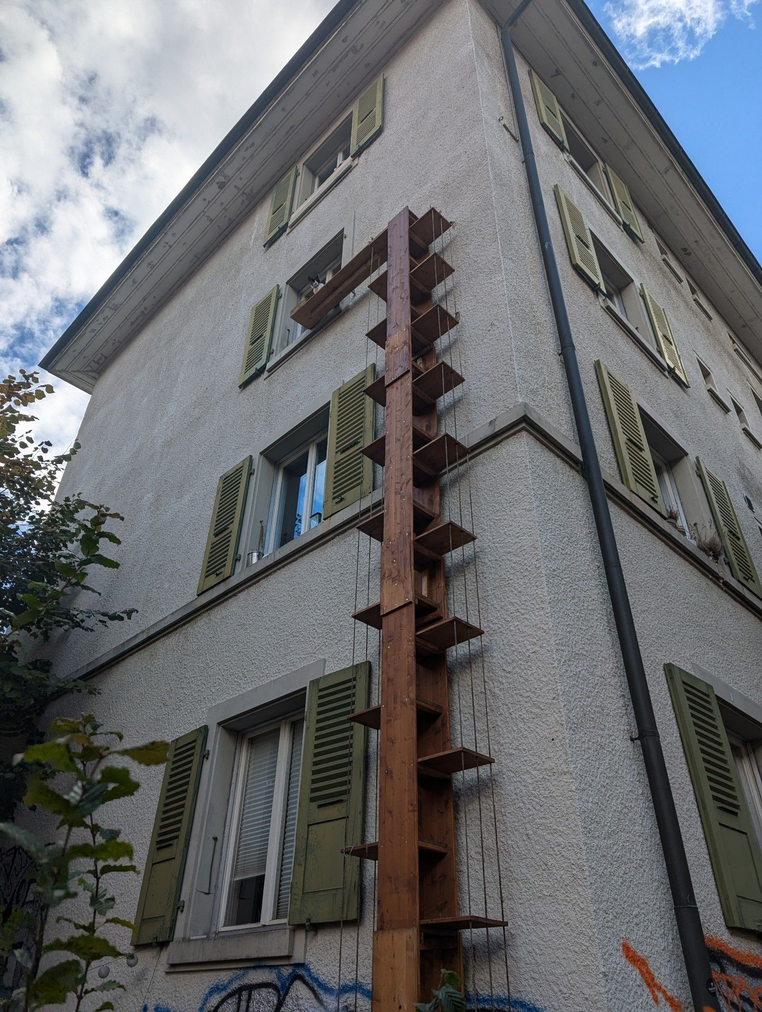 An old European multi-story townhouse. A wooden vertical staircase has been built on the side of the building allowing cats to climb up to the 3rd floor window. A cat is sitting on the top platform in front of the window looking down at the street.