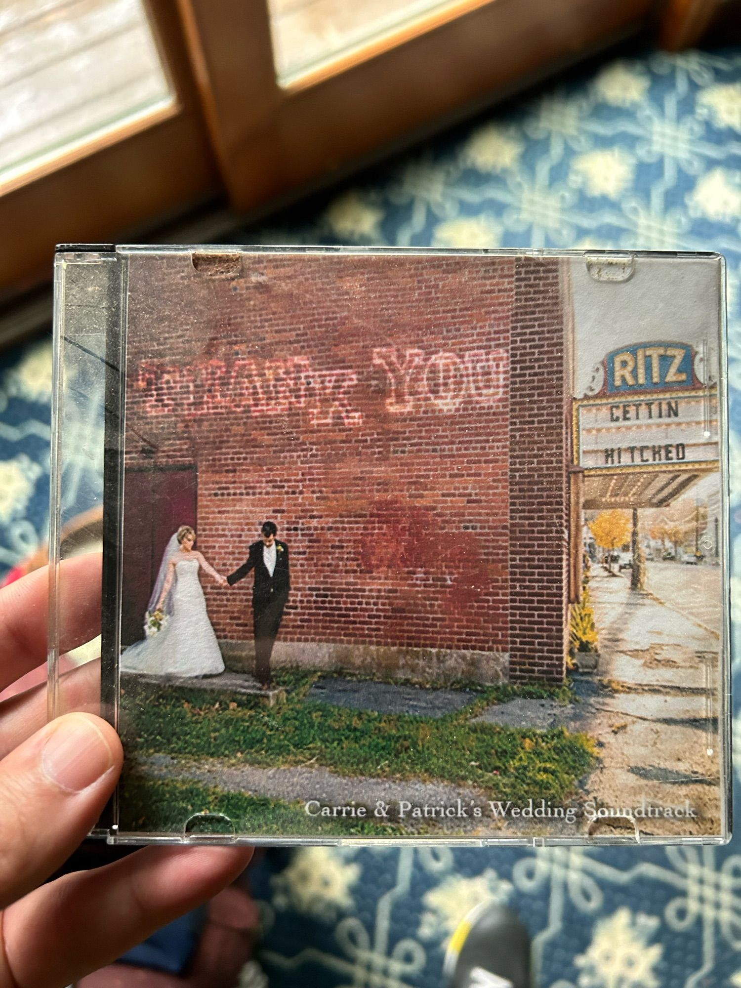 A man in a tux and woman in a wedding dress walk in front of a brick wall with thank you written on it. It’s the exterior wall of a theater and around the corner a historic marquee is visible for the ritz, and the show name is “gettin hitched”