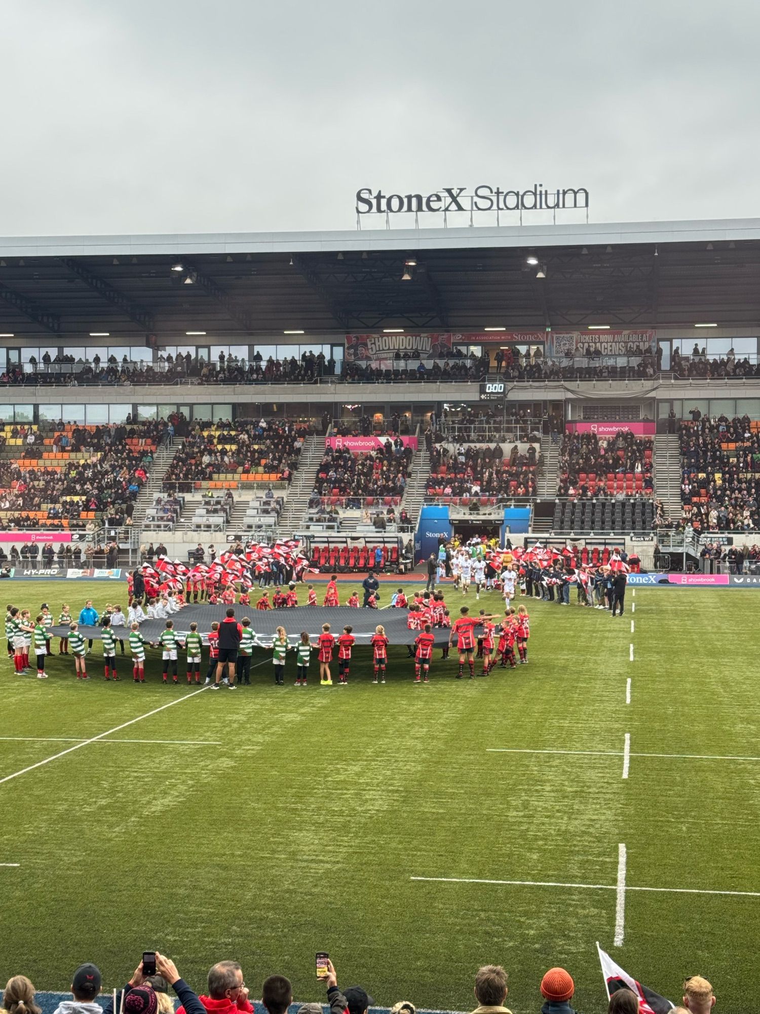 Scene of the pitch as the Exeter Chiefs team emerges from the tunnel. Lots of flags and a giant shirt held by dozens of fans.