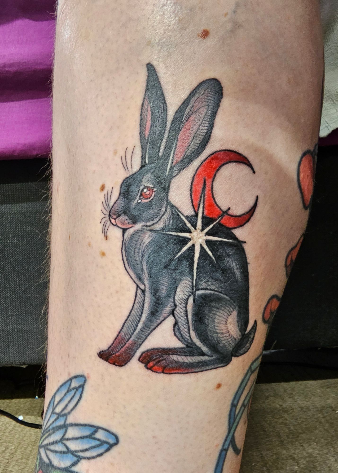 Leg tattoo. Side view of a black hare with red eyes sitting on its haunches. A white star is in front of its shoulder with a red crescent moon behind it