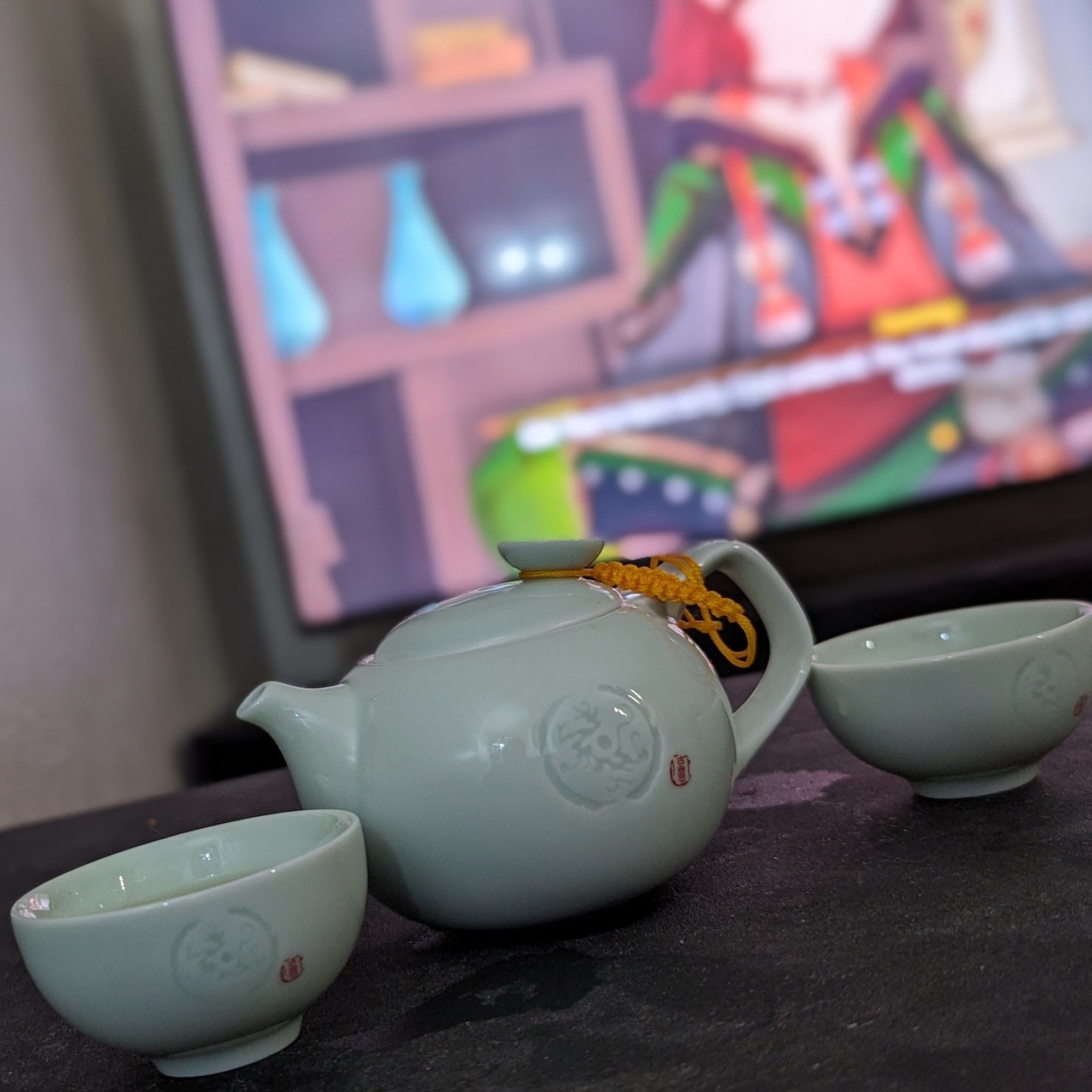 Celadon porcelain tea set in the foreground; a screen with Genshin impact blurred out in the background due to focal effects.