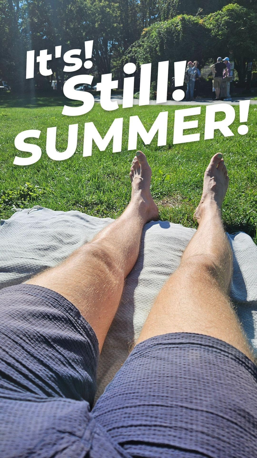 POV photo of my legs in navy shorts, no shoes, lying on a blanket in the grass in a park. Text is overlaid that says "It's! Still! SUMMER!"