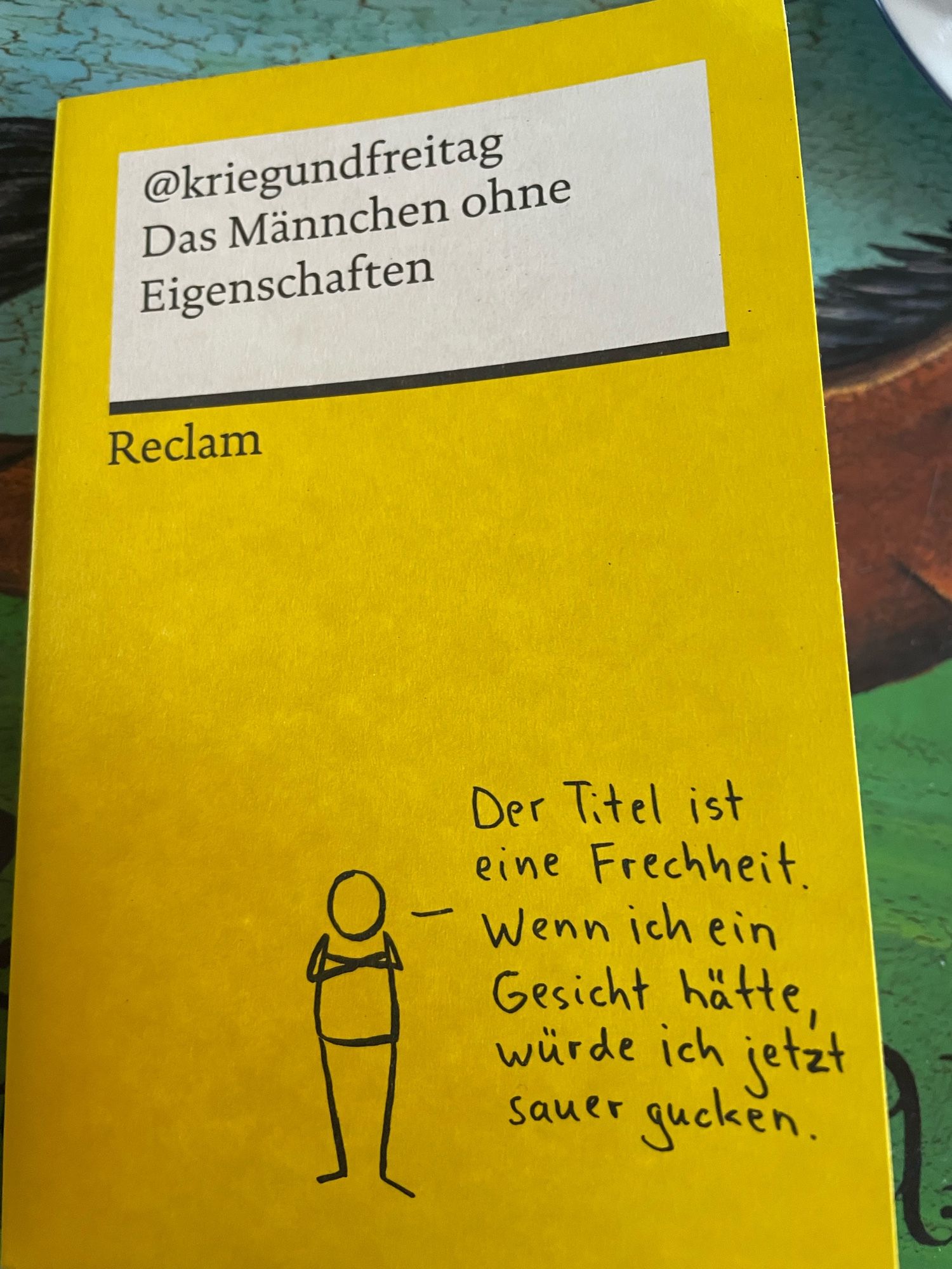 Titelbild des gelben Reclamheftes von @kriegundfreitag. Der Titel ist Das Männchen ohne Eigenschaften
Unten steht ein gesichtsloses Männchen mit verschränkten Armen. Daneben steht: Der Titel ist eine Frechheit. Wenn ich ein Gesicht hätte, würde ich jetzt sauer gucken.