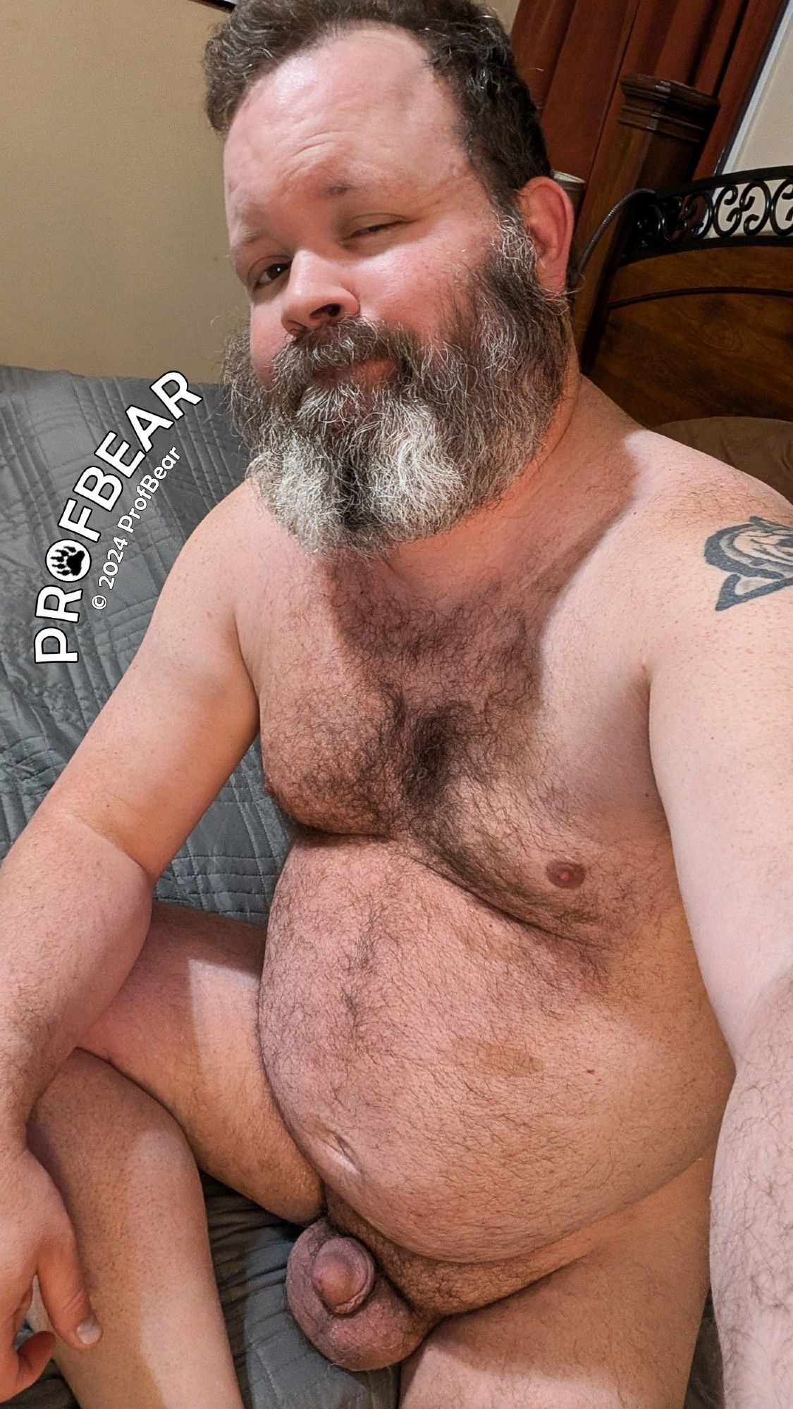 A naked, bearded, CIS man sitting in bed.