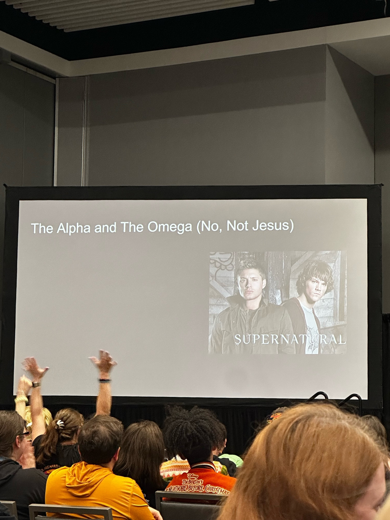 A room with people sitting in chairs and a projection reading "The Alpha and The Omega (No, Not Jesus)" featuring an image of actors Jared Padalecki and Jensen Ackles with the text "SUPERNATURAL" written underneath. 