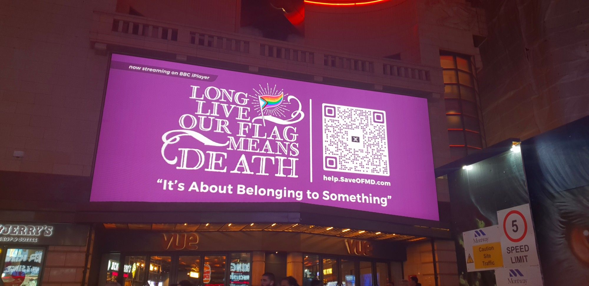 Clear shot in front of the billboard, which sits above the entrance to the Vue Cinema. The billboard says 'Long Live Our Flag Means Death' with a Progress Pride flag and a QR code with 'help.save.ofmd' underneath it. At the top it says 'streaming now on BBC iPlayer', and on the bottom it says 'it's about belonging to something'.