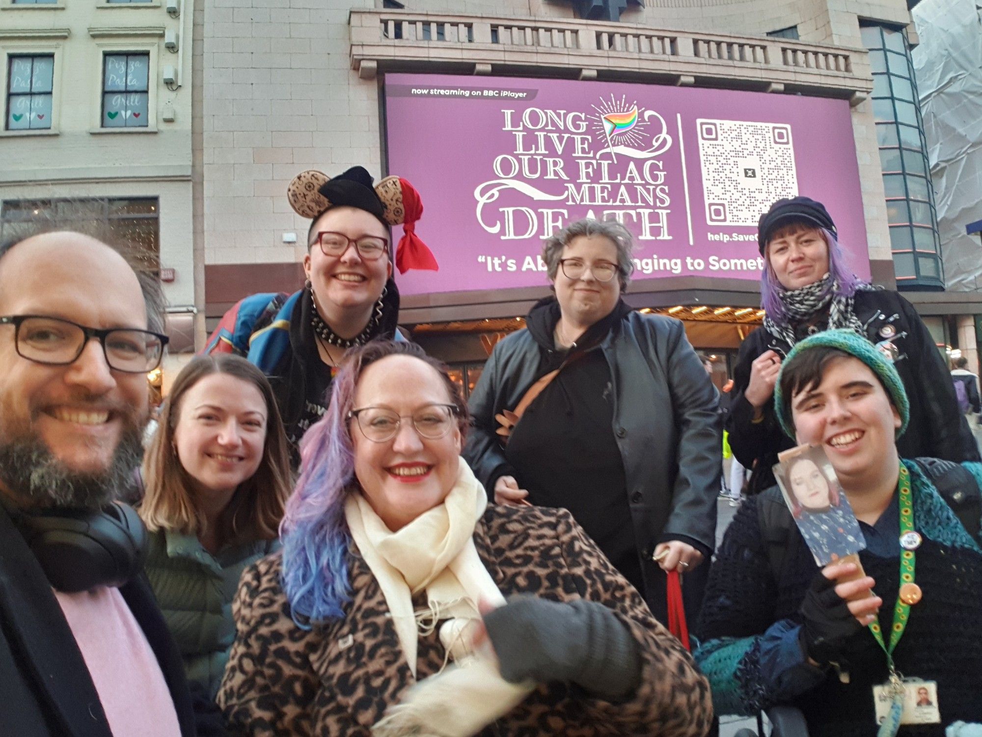 A photo of a group of smiling Our Flag Means Death fans in front of the London billboard.