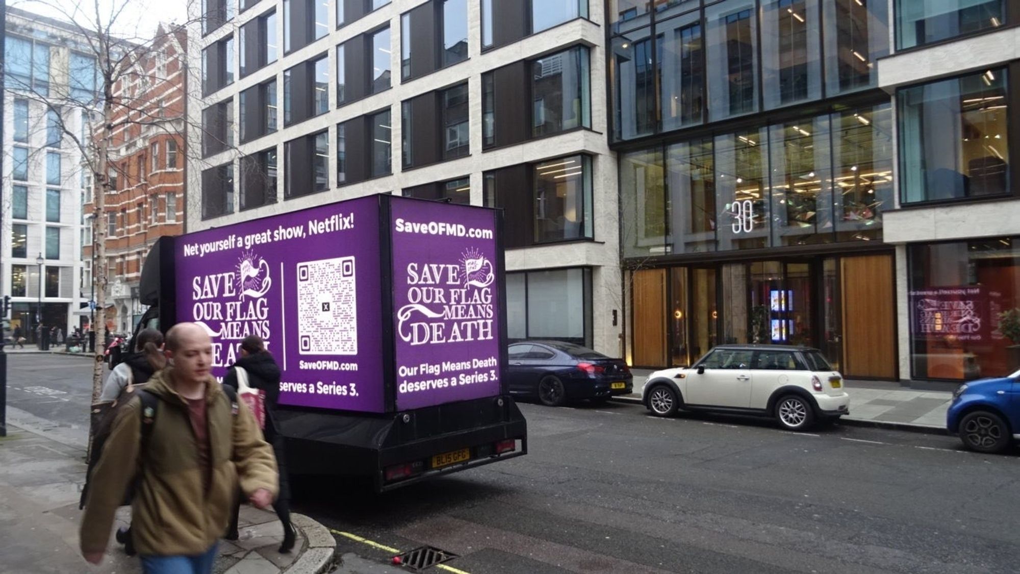 A busy London street. There is an LED advertising truck with a familiar and beautiful purple design. It has white text that reads 'Net yourself a great show, Netflix! Save Our Flag Means Death. Our Flag Means Death Deserves a Series 3.' There is a small flag above 'Flag' in the title that says '# Save O.F.M.D.' . There is a QR code to the right of the text.