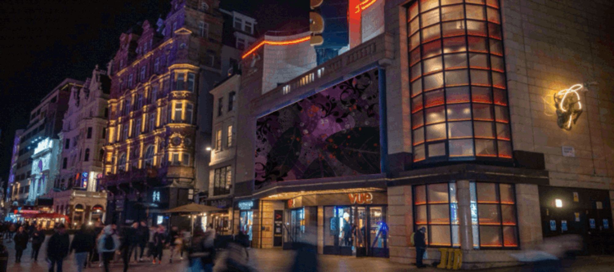 The Vue Cinema in Leicester Square at night. An animated gif has been edited onto the billboard above the cinema. The details in the animation are obscured by swirls and other decorative features in purple and black; only faint outlines of buildings and maps can be seen.