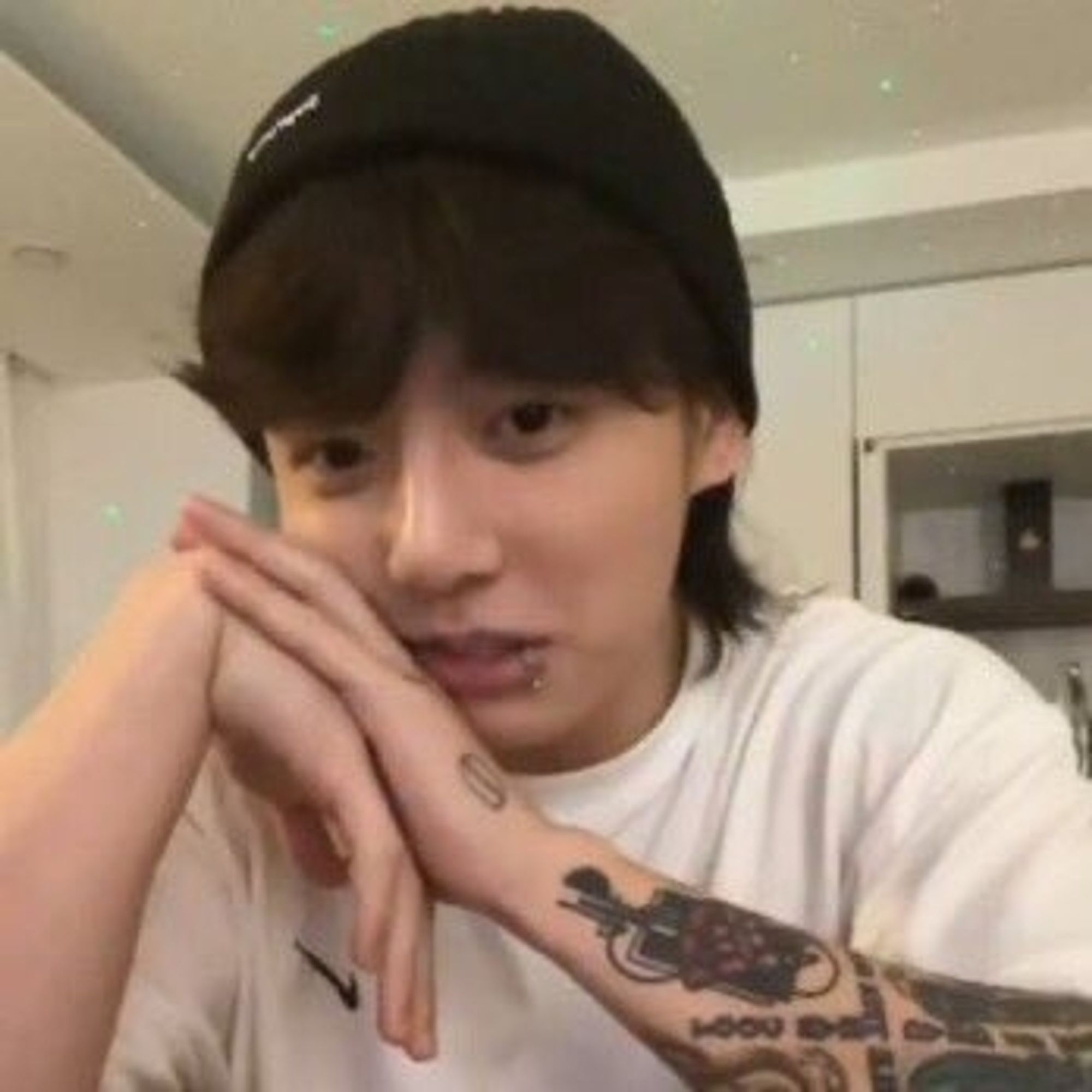 Jeon Jungkook looking all nice and soft with a plain white t-shirt and a black beanie. His face rests on his hands, tattoos partially visible as he stares into the camera mid-setence, a neutral, perhaps a little amused, expression on his face.