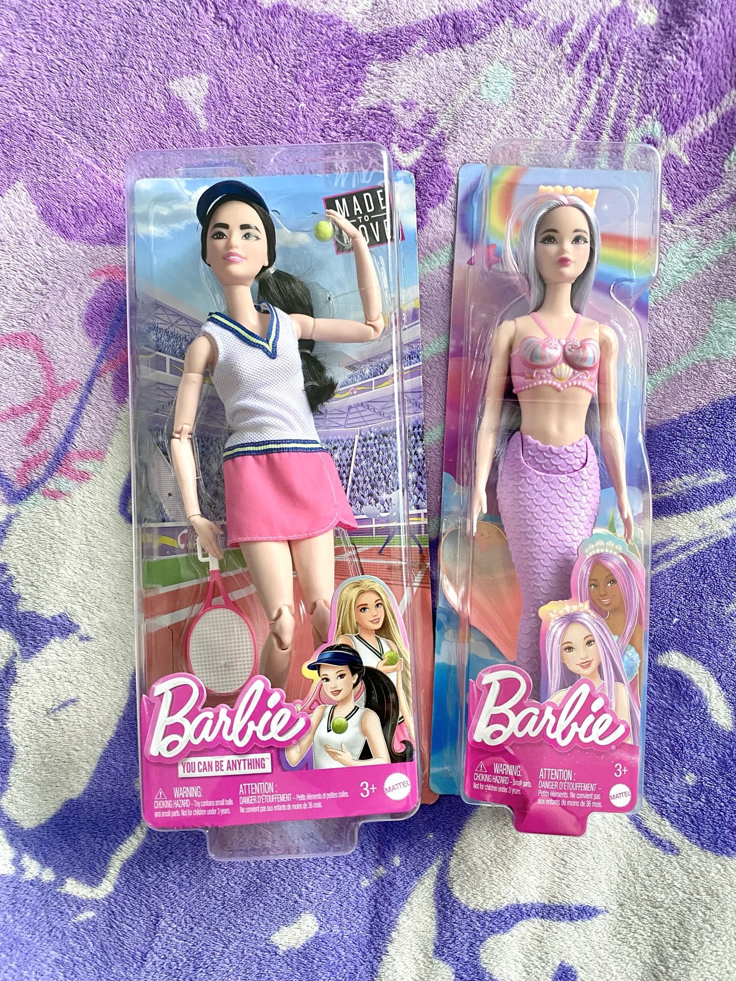 Photo of two Barbies, new in their boxes and laying on Mystic’s bed. On the left is the Barbie known as the “Made to Move Career Tennis Player,” (Product Code HKT73) and on the right is the Barbie known as the “closed-mouth Odile Mermaid.” (Product Code HRR06)