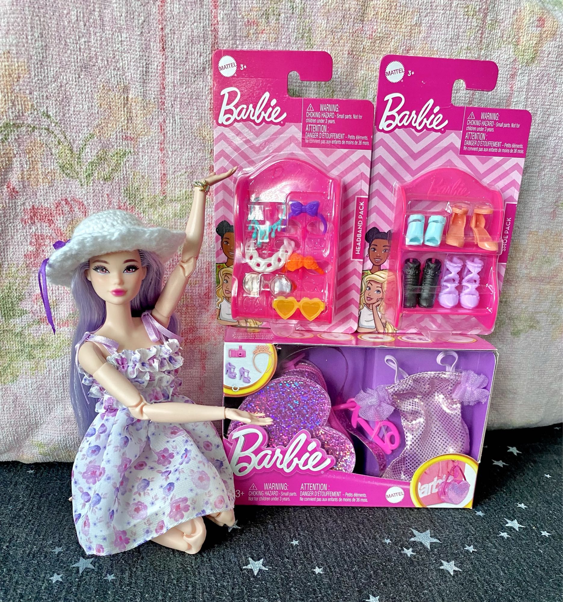 Photo of a Barbie doll* next to three Barbie accessory packs, in front of a faded floral pink backdrop. The doll is posed as sitting her on knees with her arms in a “ta-da, look at this!” Pose towards the accessories. 

Two small accessory packs—one for headbands/glasses/necklaces, one for shoes—sit atop the third, a proper Barbie fashion pack simply known as “Birthday Themed.” 

*The Barbie, named “Esmé,” to Mystic, is a hybrid made from the body of “Made to Move Career Tennis Player” (Product Code HKT73) and the head of a lavender-haired mermaid doll (Product Code HRR06). She is wearing the dress and bracelet from the “@BarbieStyle Spring Date” fashion pack and a white crochet sun hat with a decorative purple ribbon.