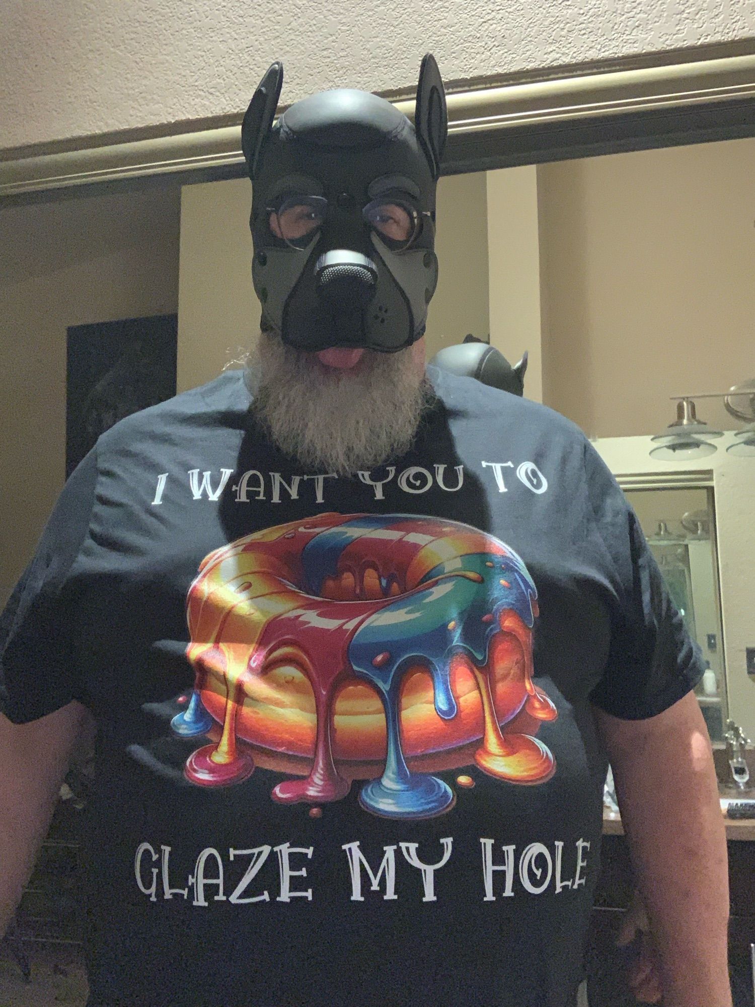 Chubby pup in a black pup hood with a gray muzzle. He’s wearing a black t-shirt with a donut centered on the shirt with a rainbow colored glaze dripping down the sides of the donut. The t-shirt says, “I want you to glaze my hole.”