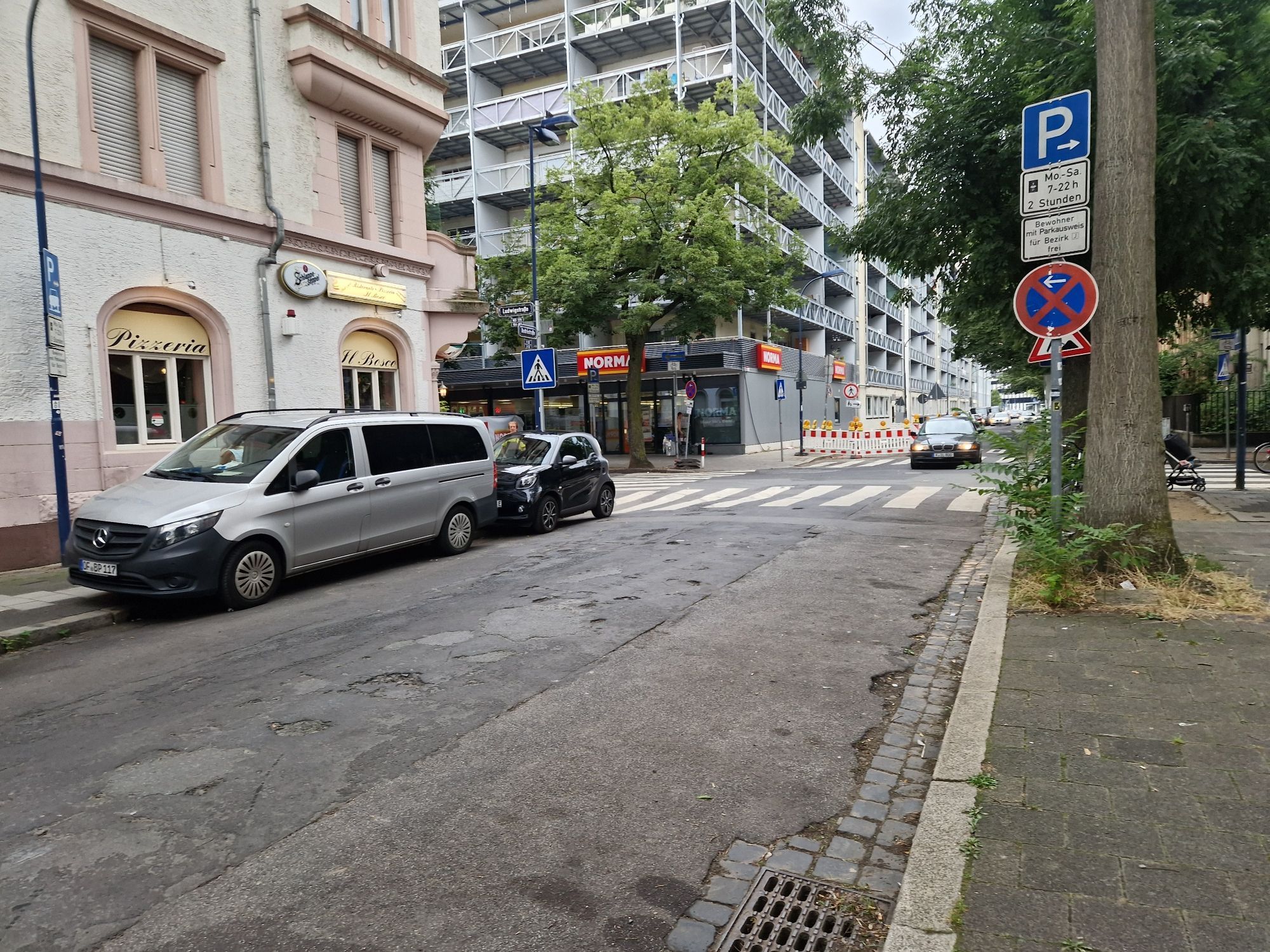 Straße mit Kreuzung, Zebrastreifen und falsch geparkten Fahrzeugen halb auf den Gehsteig aufgesetzt