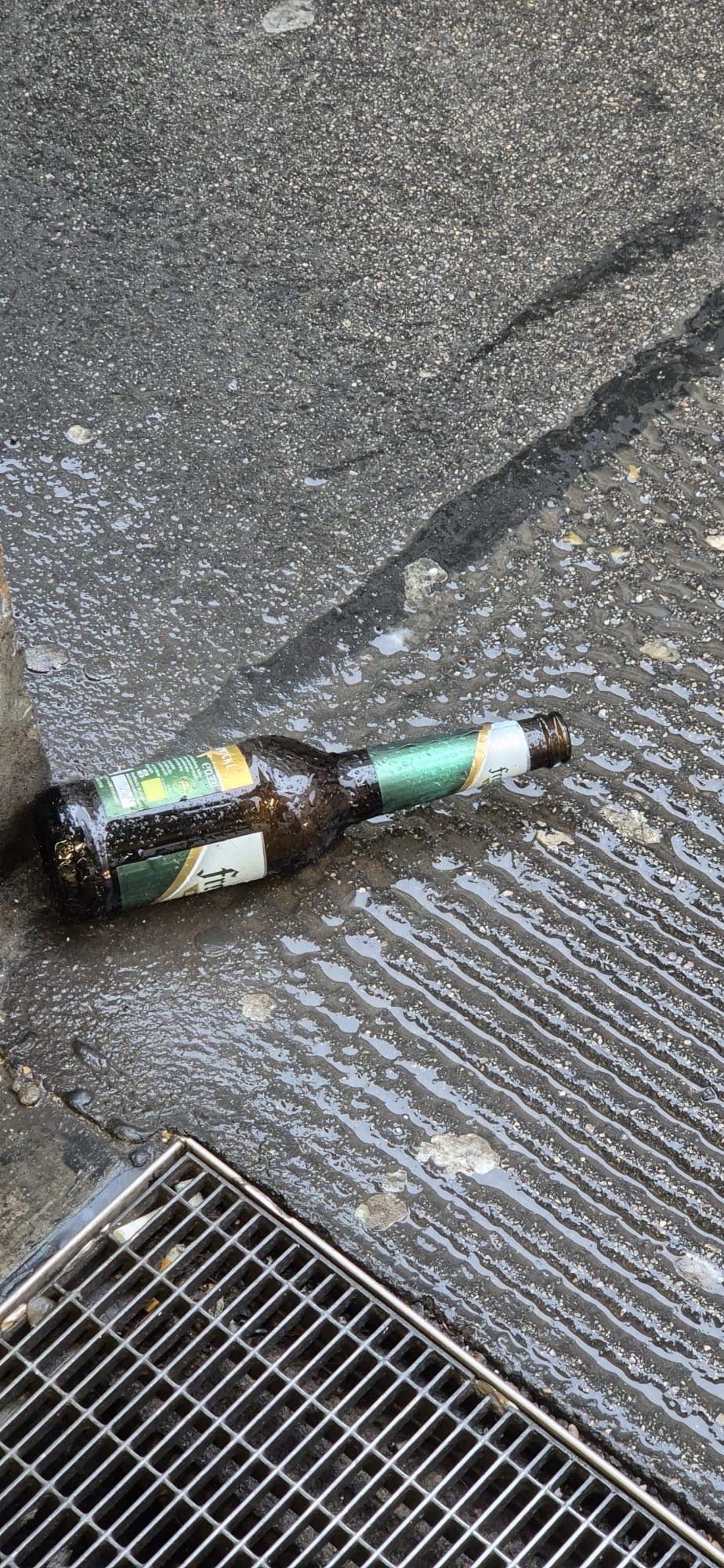 Braune Bierflasche mit grünem Etikett liegt auf nassem Granitboden, am unteren Bild sieht man einGitter