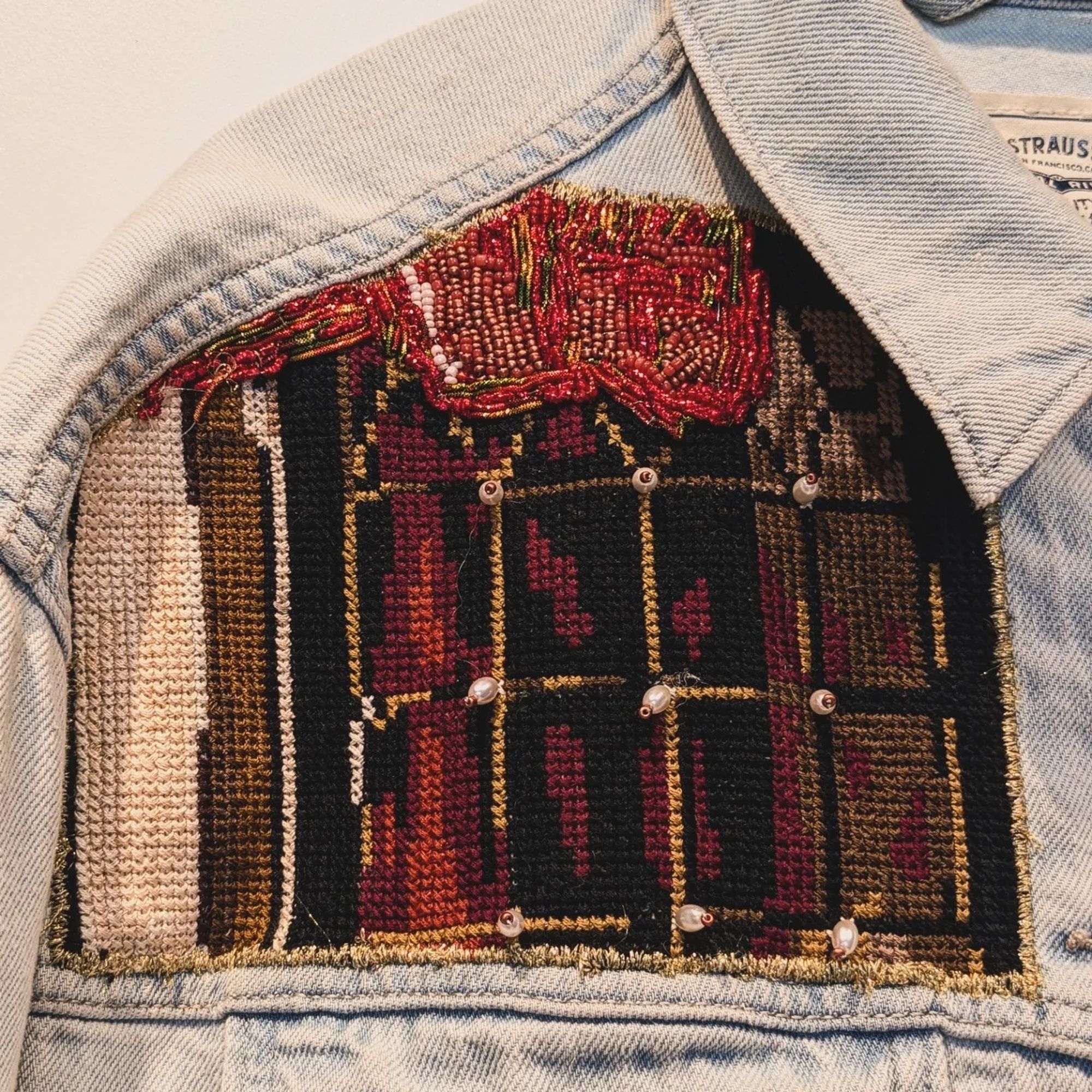 An embroidered patch on the left breast of a light jean jacket. It shows a paned window with a length of red cloth hanging off it. The window is rendered in cross stitch and the cloth in bead work.