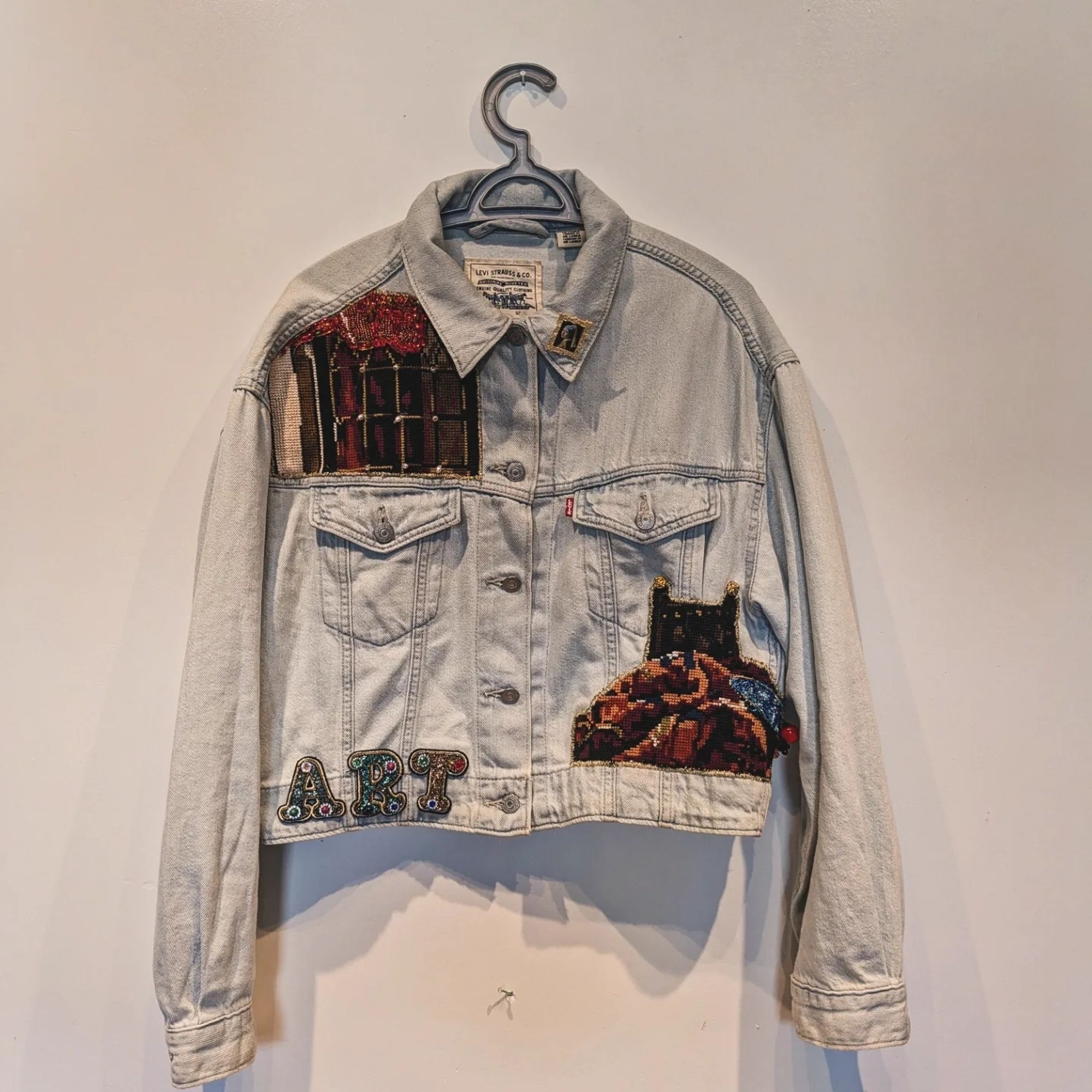 A light blue jean jacket hanging up against a white wall. There are embroidered and beaded sections in the upper left and lower right sections of the body. On the lower left section is the word ART spelled out in bedazzled letters.