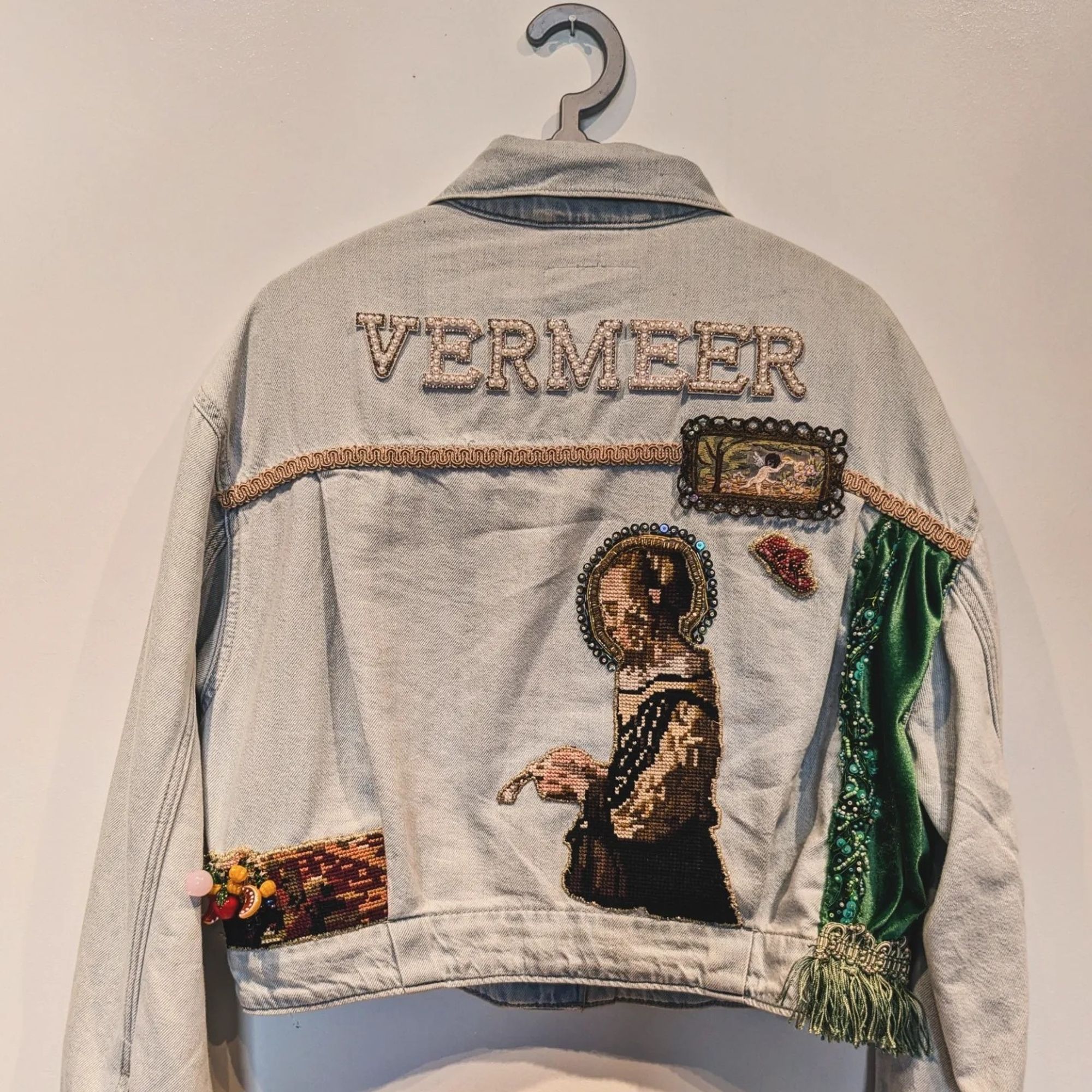 A light jean jacket hanging up against a white wall. Across the shoulders it says VERMEER in bedazzled letters. There's a patch of an old timey lady reading a letter. She has a halo around her. There are also patches of a cherub in a frame and a devil mask. The right hand side is covered in embroidered green velvet and fringe.