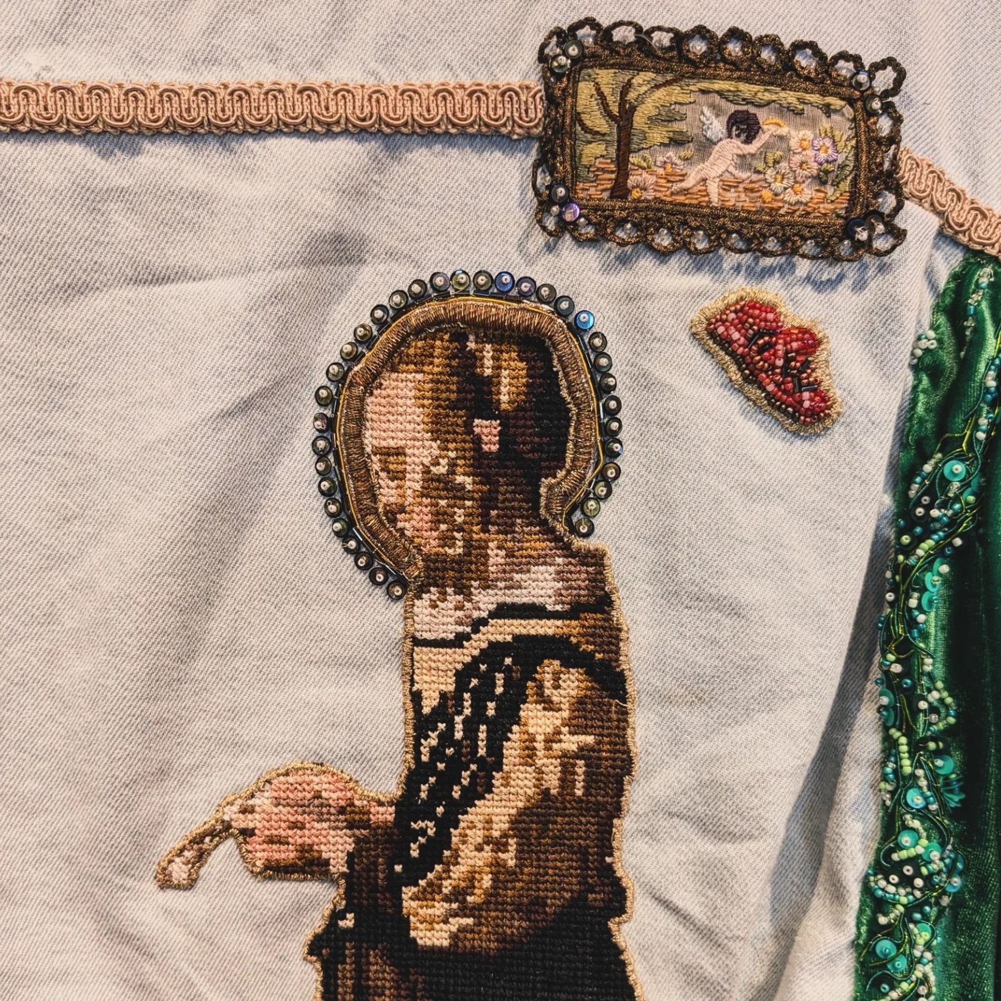 Close up of three patches on the back of a light blue jean jacket. There's a cross stitch of an old timey lady reading a letter. She has a bead and hold bullion halo. There's a bead work devil face. And an embroidered patch of a cherub in a faux frame. To the right is green embroidery and bullion work.