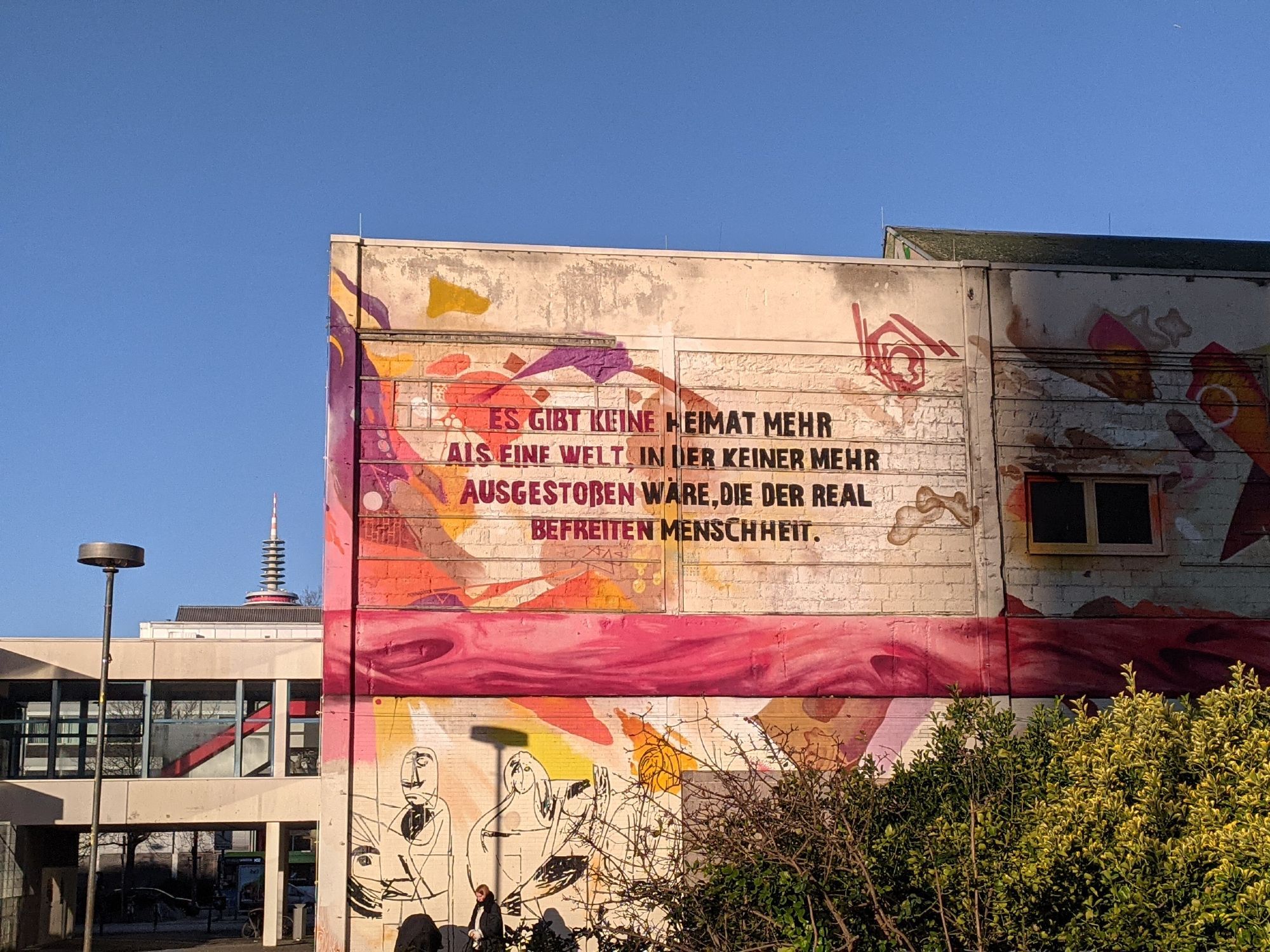 Es gibt keine Heimat mehr als eine Welt, in der keiner mehr ausgestoßen wäre, die der real befreiten Menschheit - Zitat von Theodor Wiesengrund Adorno in Wandschrift hoch über dem Campus Bockenheim am Gebäude Labsaal, zu letzt eine Not-Unterkunft für Geflüchte.
Blauer Winterhimmel, im Hintergrund die Spitze des Ginnheimer Spargel, dem Fernsehturm