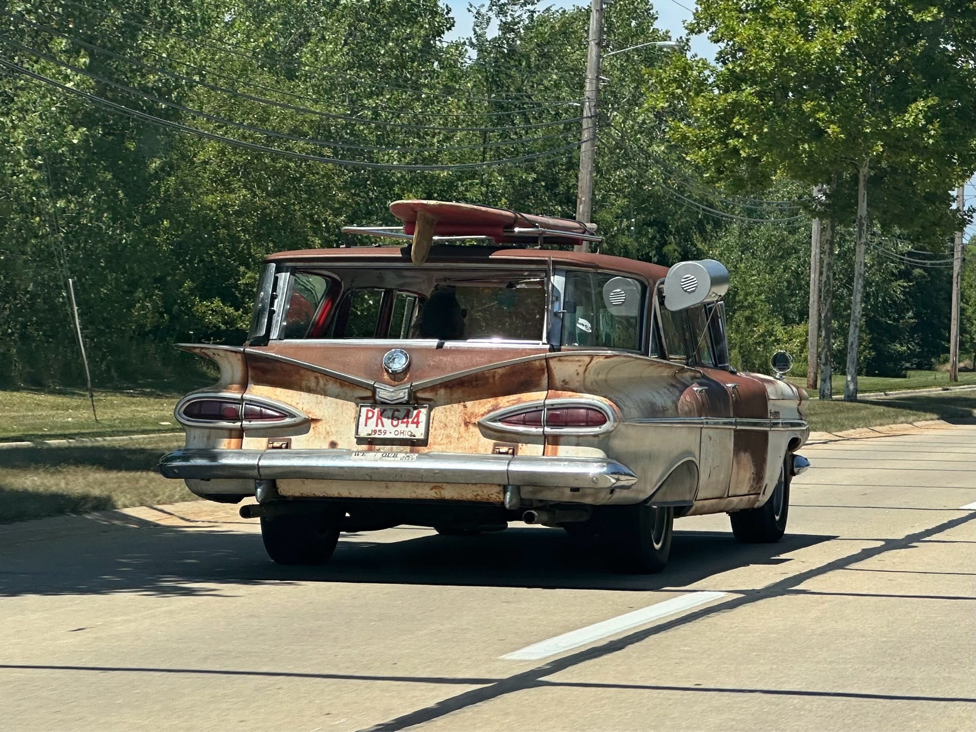 A rear view of a late 50s Chevy Kingswood station wagon, showing heavy patina. There’s a surfboard mounted on the roof rack.