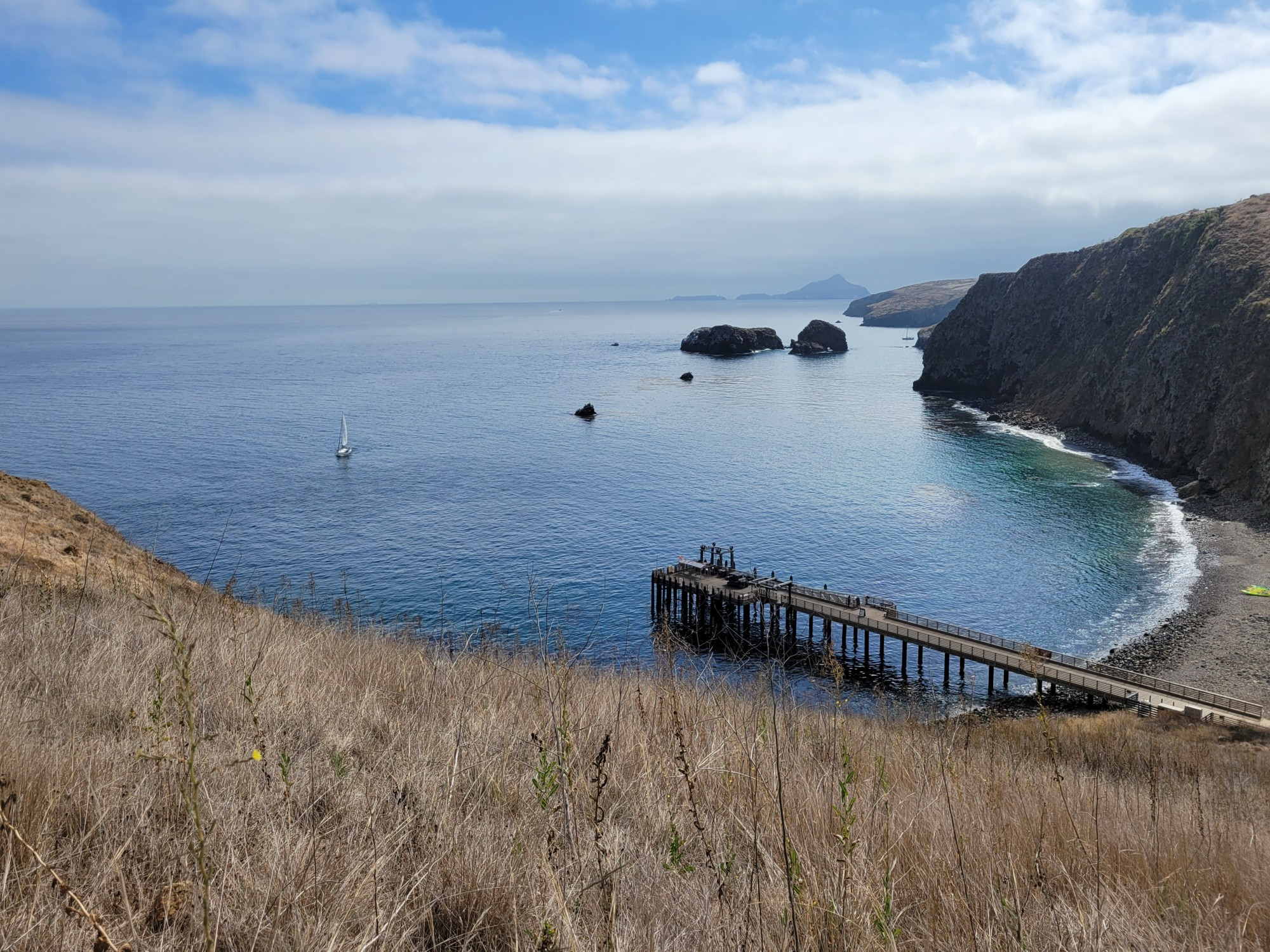 Looking down over the pier, the beach, and the ocean. With portions of the island and another island in the distance. Dead grass/brush in the foreground. A sailboat is in the water.