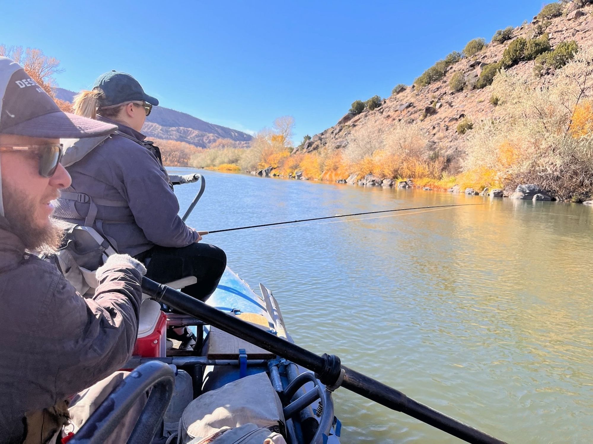 Myself in the bow of the raft and our guide sitting mud boat with oars. My flies are in the water and the top of the rod is pointing at the riparian area that is in its autumn oranges and yellows.  The guide is in wool gloves indicating just how chilly it was.