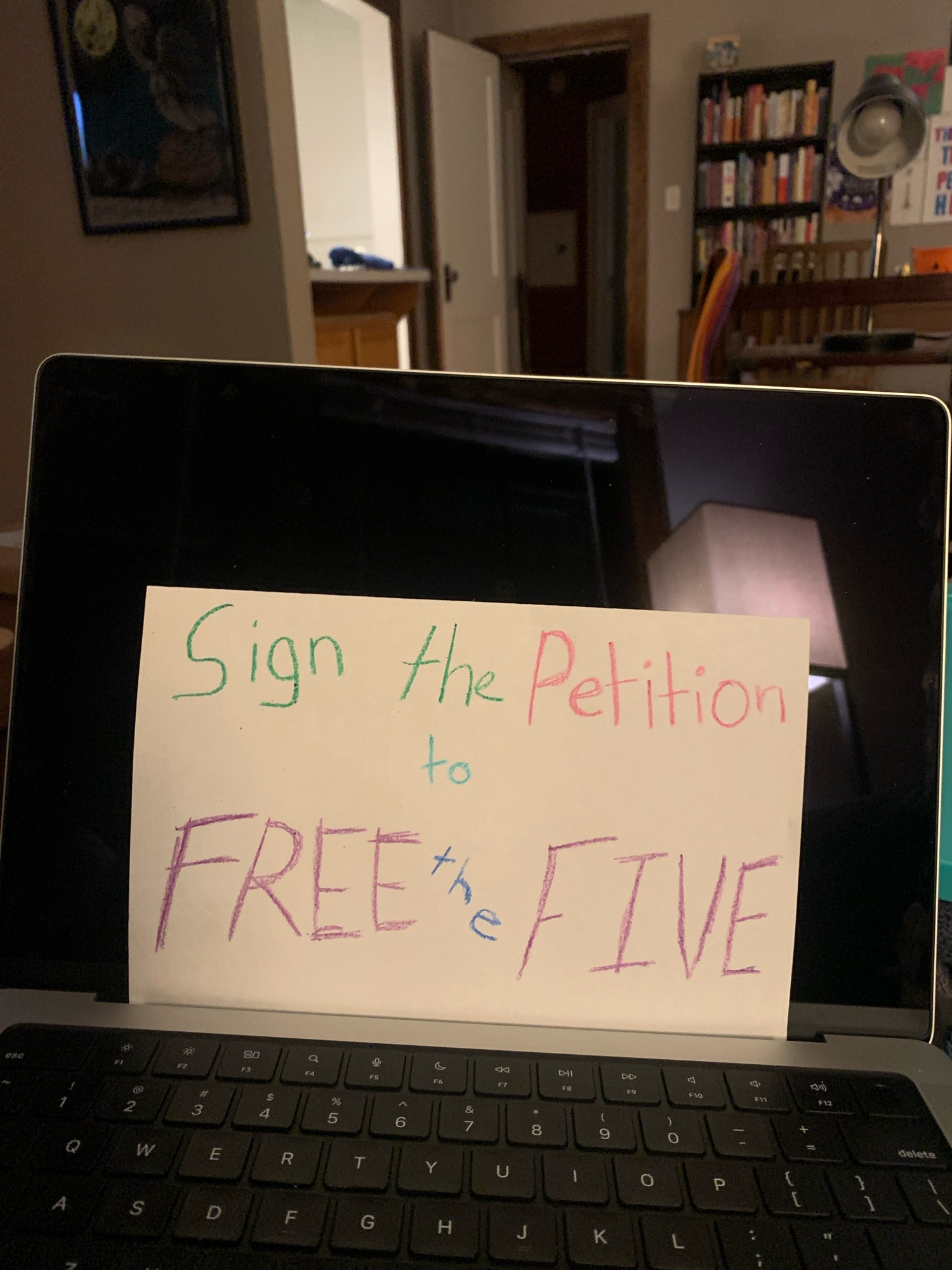 A small sheet of paper rests against a blank computer screen. On the sheet, written in crayon, it reads “Sign the petition to FREE the FIVE”